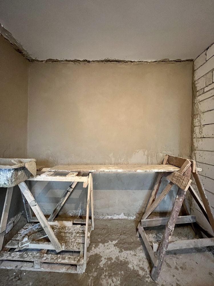 Świeżo otynkowana ściana w pomieszczeniu, widoczne drewniane rusztowania i narzędzia murarskie. Fragment ściany z cegły. Szare tynki na ścianach i suficie.