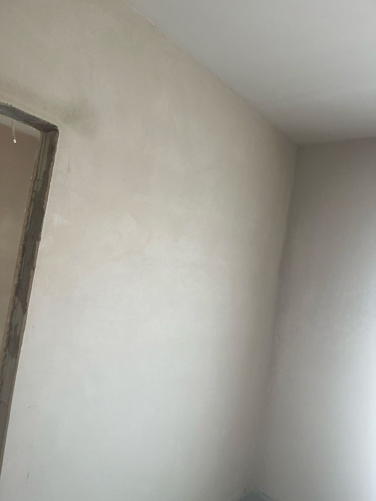 Świeżo położona, jasna gładź na ścianie w rogu pokoju, widoczna surowa ościeżnica drzwiowa. Delikatne światło podkreśla fakturę. Minimalistyczne wykończenie wnętrza.