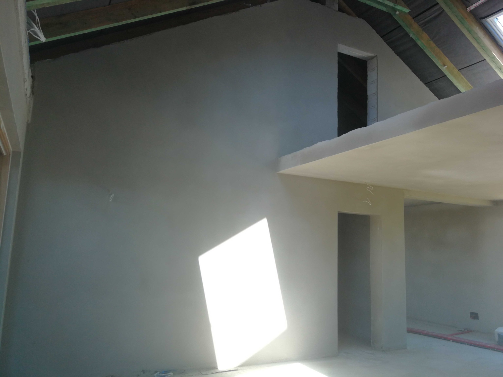 Świeżo otynkowane wnętrze poddasza z widoczną konstrukcją dachu i wpadającym światłem słonecznym, ukazujące gładkie, szare ściany przed dalszym wykończeniem.