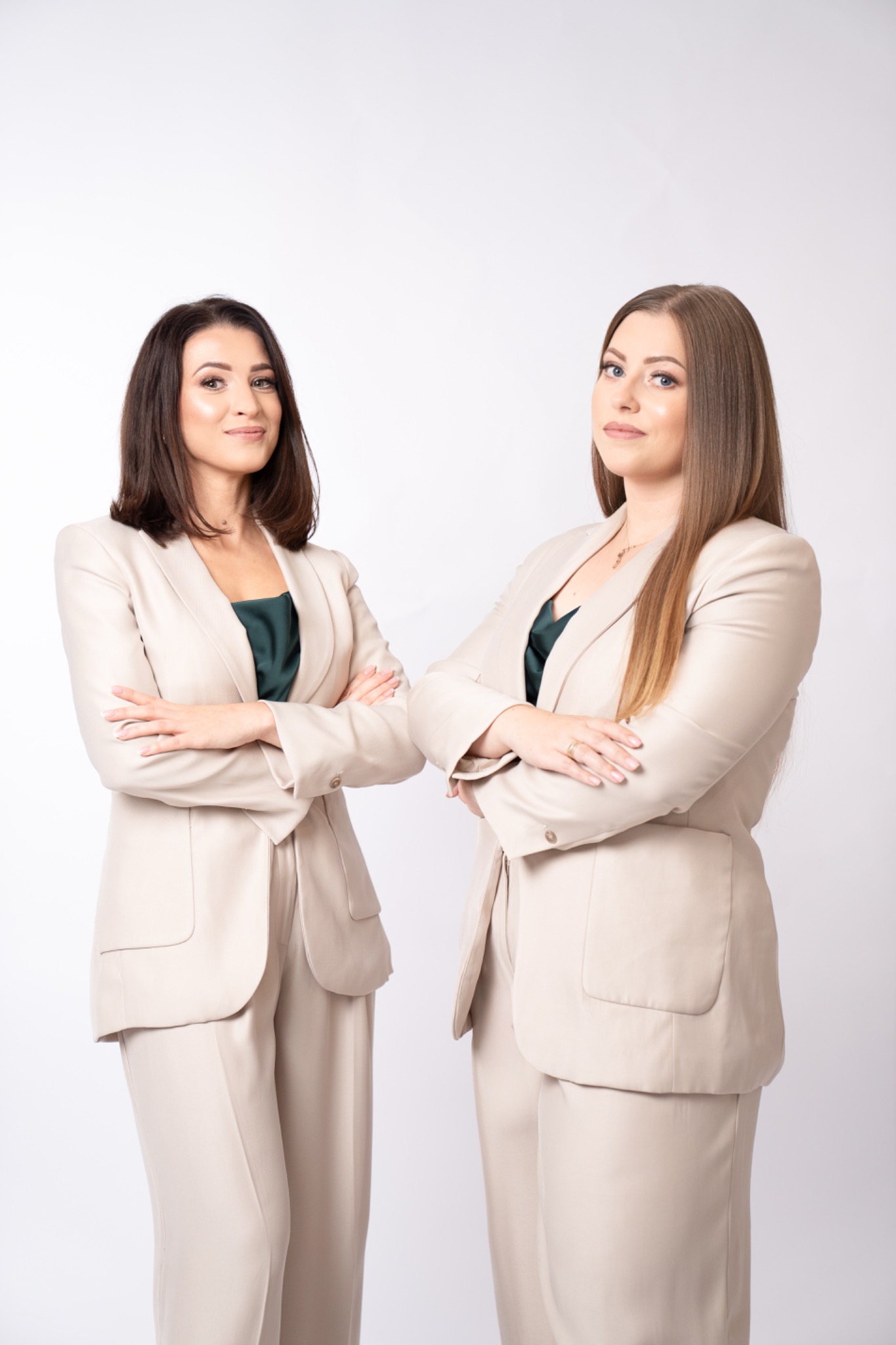 Dwie uśmiechnięte kobiety w beżowych garniturach, stojące na białym tle z założonymi rękami. Profesjonalny wizerunek zespołu, budzący zaufanie.