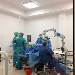 Medica Vision Warszawa - Okulistyka - Optyk - Sala operacyjna z zespołem medycznym w trakcie zabiegu. Widoczny sprzęt specjalistyczny, oświetlenie i personel w strojach ochronnych. Czerwone wiaderko na podłodze.