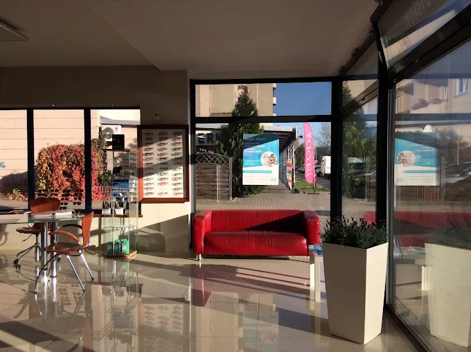 Jasne wnętrze salonu optycznego z czerwonym siedziskiem, ekspozycją okularów i widokiem na ulicę przez duże okna. Podłoga z połyskiem odbija światło.