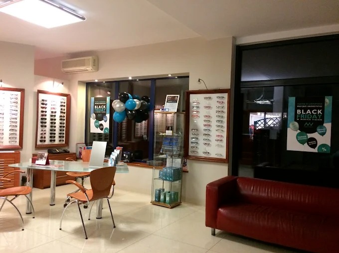 Wnętrze salonu optycznego z ekspozycją okularów w ramkach, stolikiem, krzesłami i czerwoną sofą. Dekoracja balonowa z okazji Black Friday.