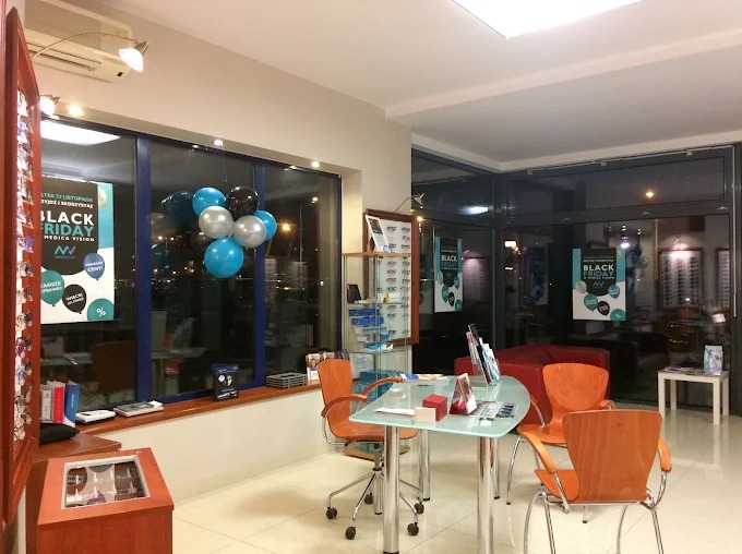 Jasne wnętrze salonu optycznego z ekspozycją okularów, stolikiem i krzesłami. Na oknach balony i plakaty informujące o promocji Black Friday.