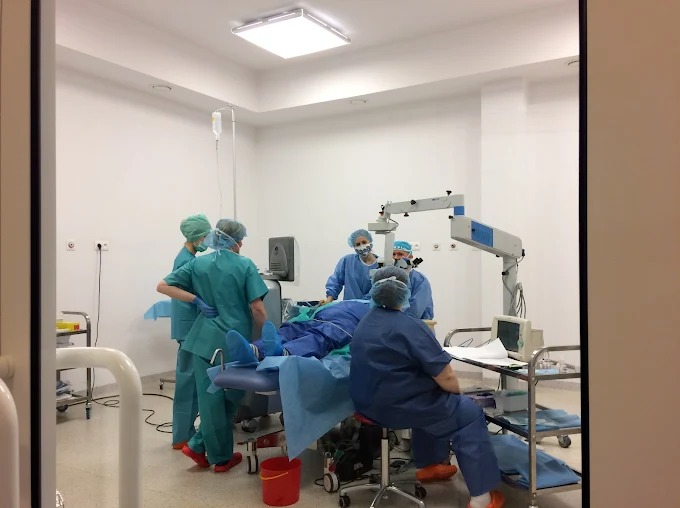 Sala operacyjna z zespołem medycznym w trakcie zabiegu. Widoczny sprzęt specjalistyczny, oświetlenie i personel w strojach ochronnych. Czerwone wiaderko na podłodze.