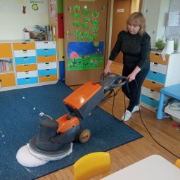 NataGroup - Kobieta czyści dywan w przedszkolu za pomocą profesjonalnej maszyny TASKI. Widoczne kolorowe szafki i dekoracje dziecięce. Podłoga drewniana, jasne oświetlenie.