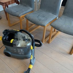 NataGroup - Ekstraktor do tapicerki Karcher Puzzi obok trzech krzeseł z plamami na siedziskach, na jasnej podłodze z paneli. Sprzęt gotowy do czyszczenia.