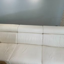 NataGroup - Fragment białej, skórzanej sofy z widocznym obszarem po czyszczeniu, ukazującym różnicę w odcieniu i czystości. Tło stanowi szara ściana i fragment ściany z białej cegły.