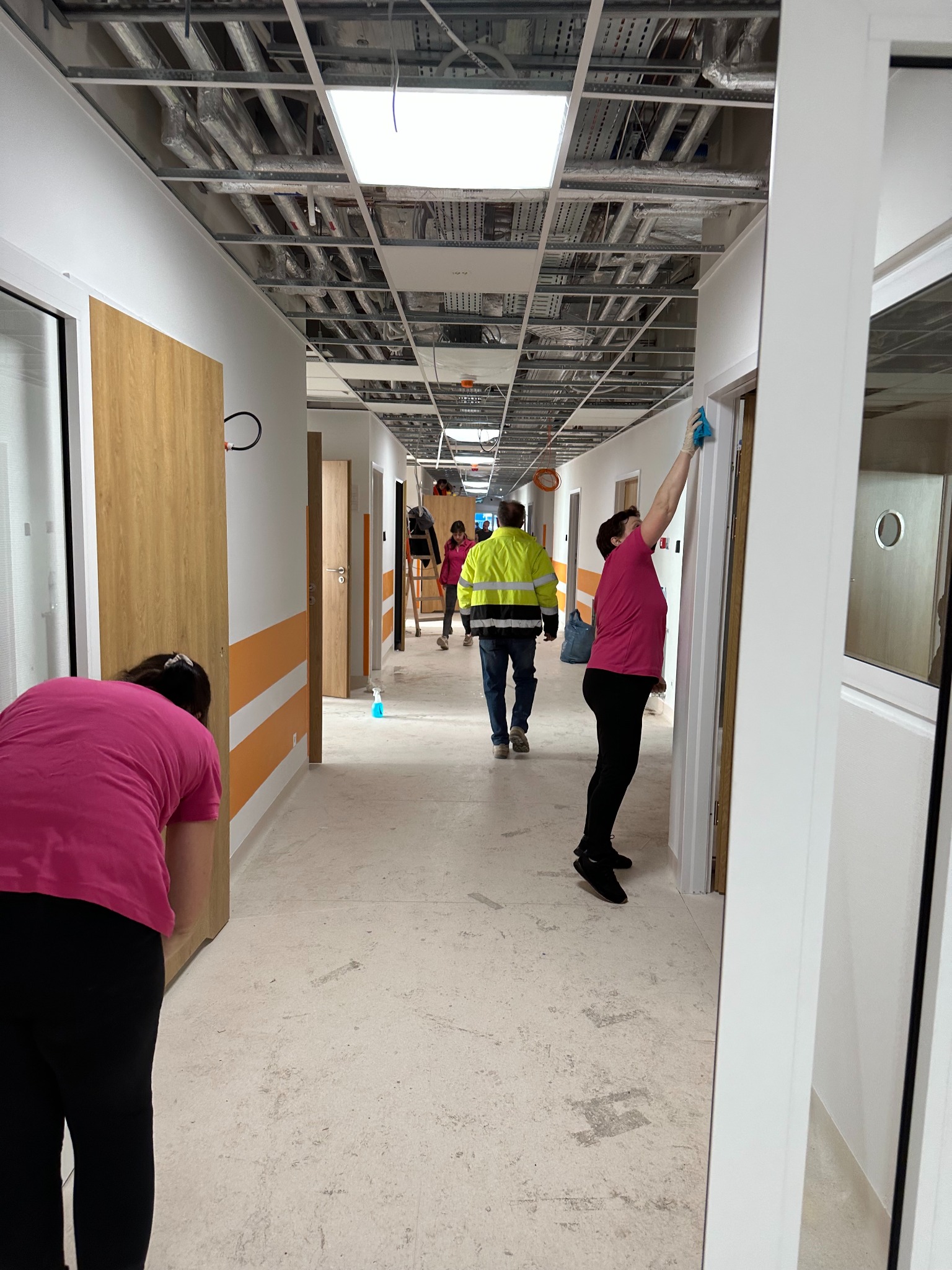 Korytarz w trakcie sprzątania po remoncie. Widoczne osoby czyszczące ściany i podłogę, butelka ze środkiem czyszczącym oraz surowy sufit z instalacjami.
