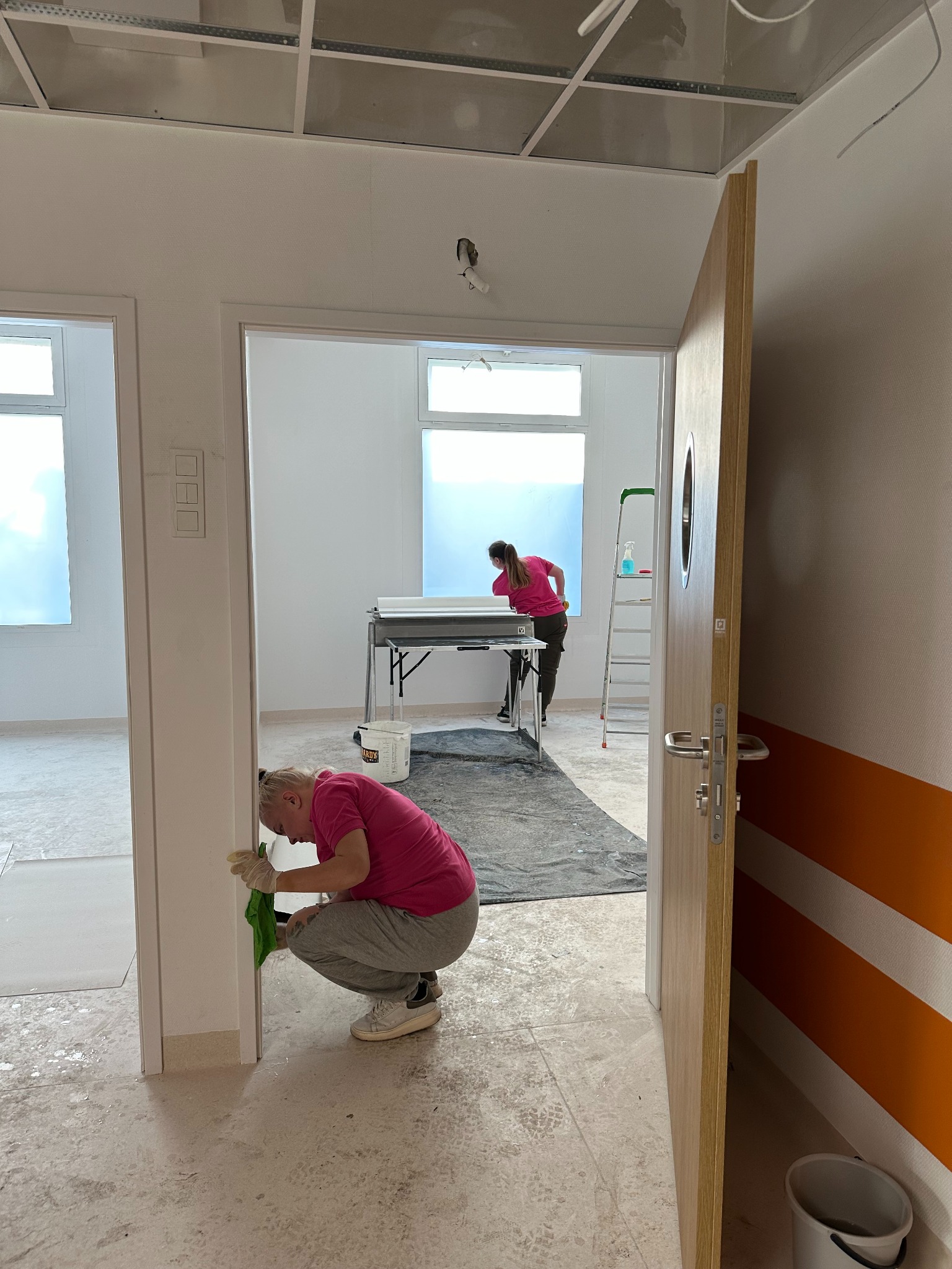 Dwie osoby w różowych koszulkach czyszczą wnętrze budynku po remoncie. Jedna osoba czyści framugę drzwi, druga okno. Widoczne wiadro, drabina i folia ochronna.