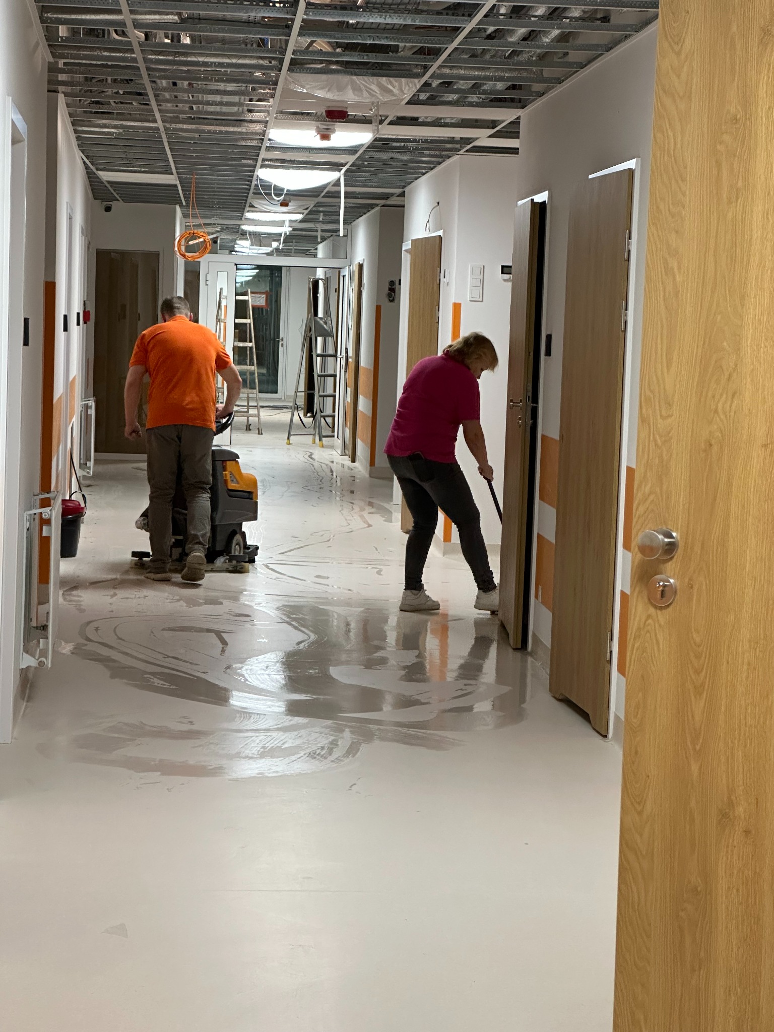 Mycie podłogi w korytarzu budynku w trakcie budowy. Dwie osoby czyszczą podłogę za pomocą maszyny i mopa. Widoczne niedokończone sufity i drzwi.