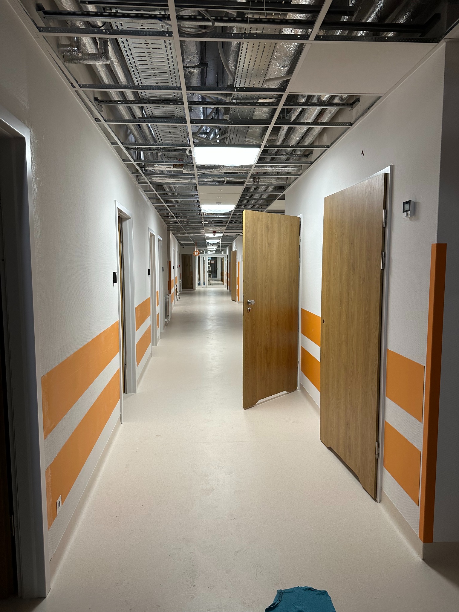 Długi korytarz w budynku w trakcie wykańczania, z widocznymi instalacjami pod sufitem, pomalowanymi ścianami z pomarańczowymi pasami i otwartymi drzwiami.