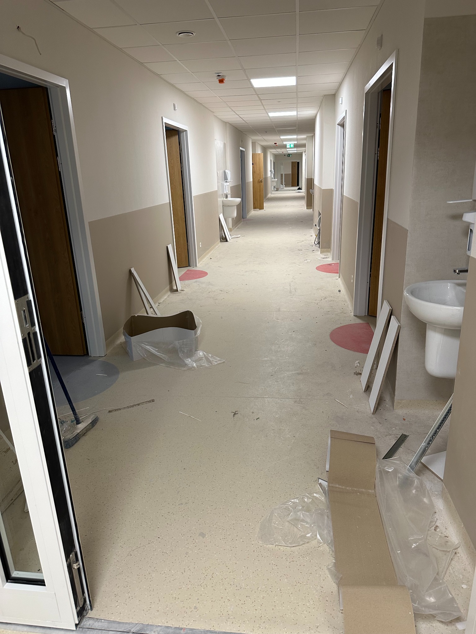 Długi korytarz w budynku po remoncie. Widoczne umywalki, drzwi, resztki materiałów budowlanych i ślady po pracach wykończeniowych na podłodze.