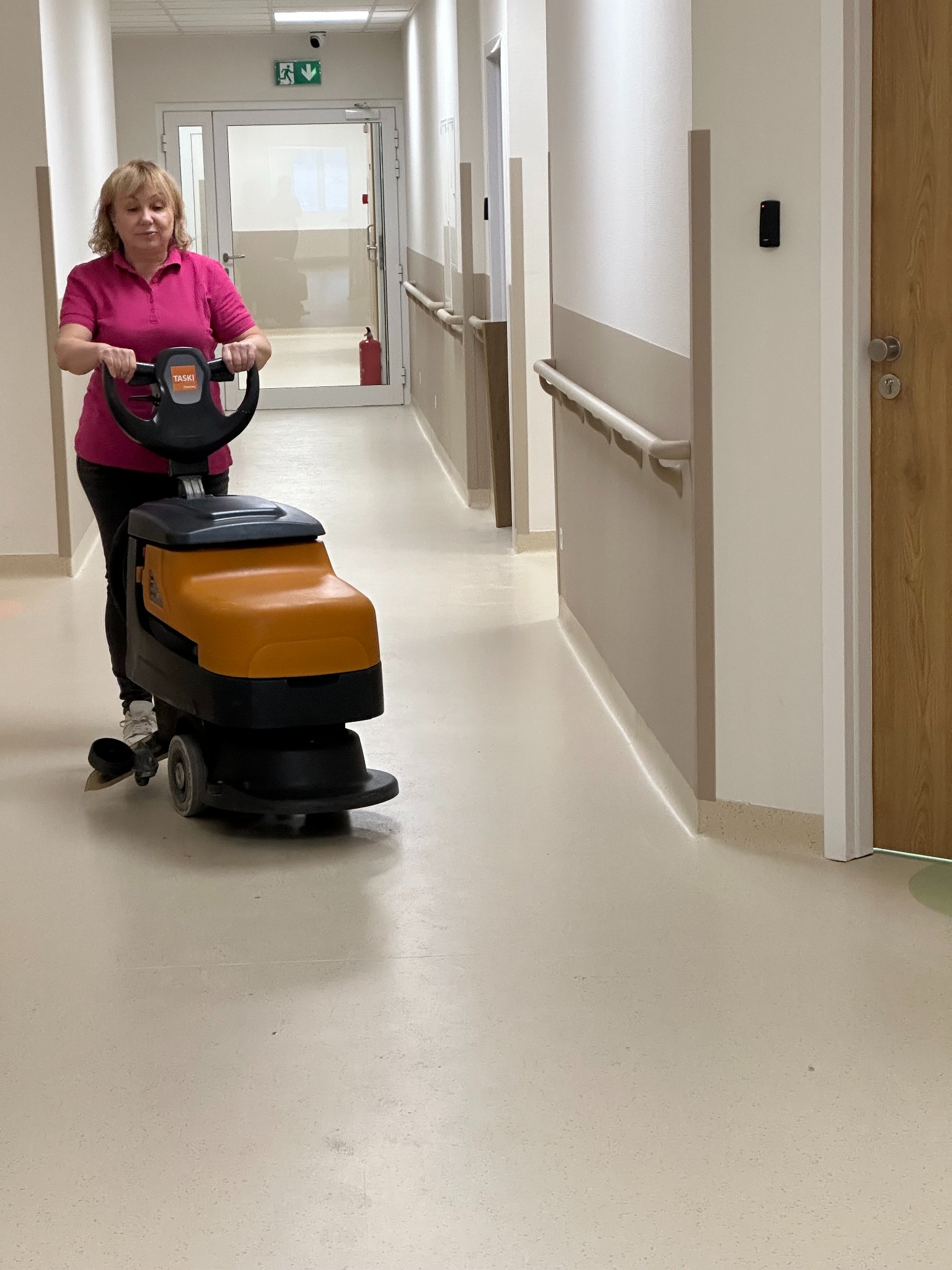 Kobieta obsługuje pomarańczową maszynę do czyszczenia podłóg w jasnym korytarzu szpitalnym z poręczami i drzwiami. Widoczny znak ewakuacyjny.