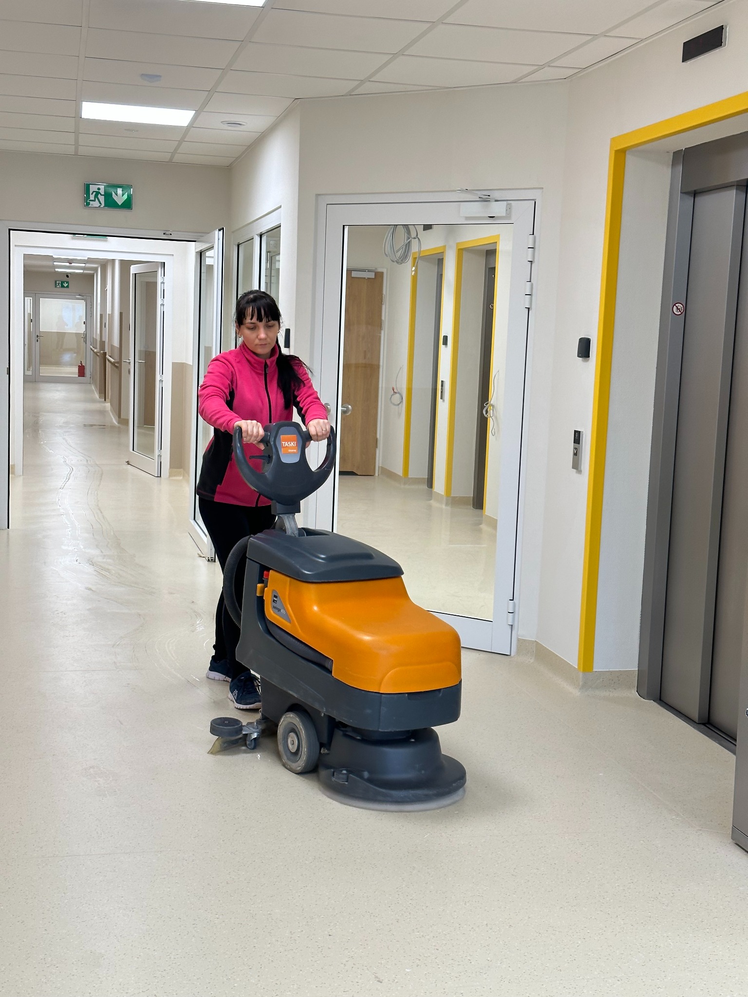 Kobieta w różowej bluzie obsługuje maszynę czyszczącą podłogę w jasnym korytarzu z żółtymi akcentami. Widoczne ślady po czyszczeniu i drzwi do windy.