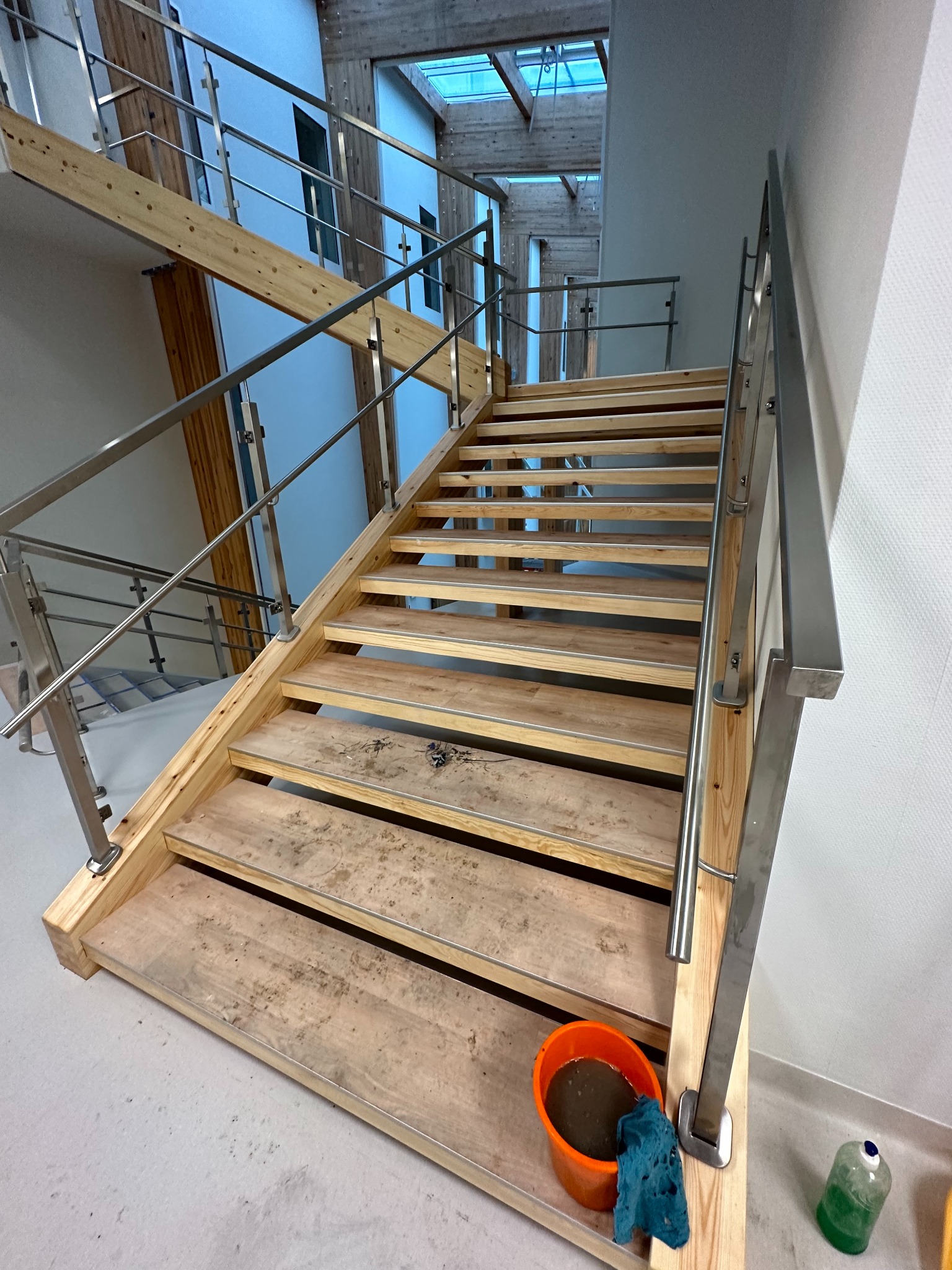 Drewniane schody z metalową balustradą w jasnym wnętrzu. Widoczne ślady po budowie, wiadro z wodą i ścierka. Ujęcie z dołu, podkreślające wysokość i konstrukcję.