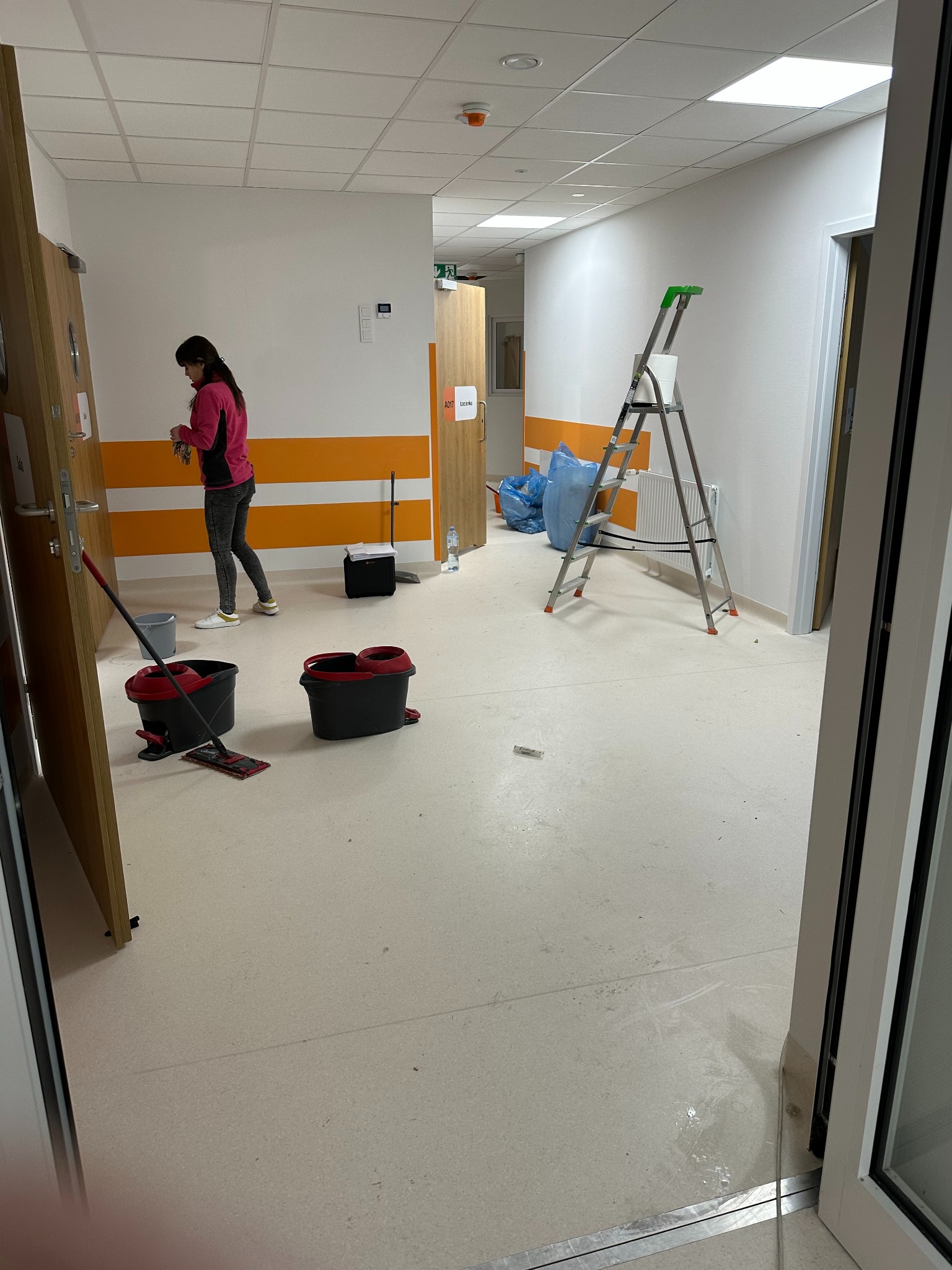 Korytarz w trakcie sprzątania: kobieta z mopem, wiadro, drabina, worki na śmieci. Widoczne mokre ślady na podłodze, pomarańczowe pasy na ścianach i biały sufit kasetonowy.