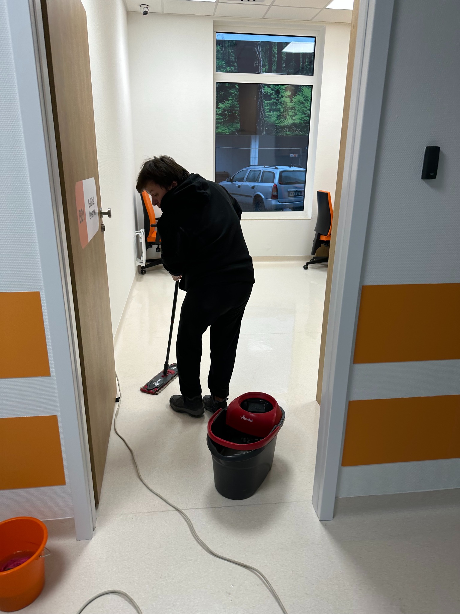 Osoba w czarnym ubraniu myje podłogę mopem w jasnym pomieszczeniu z oknem. Widoczny wózek z mopem, wiadro i kabel. Na drzwiach tabliczka z napisem 'Gabinet Lekarski'.