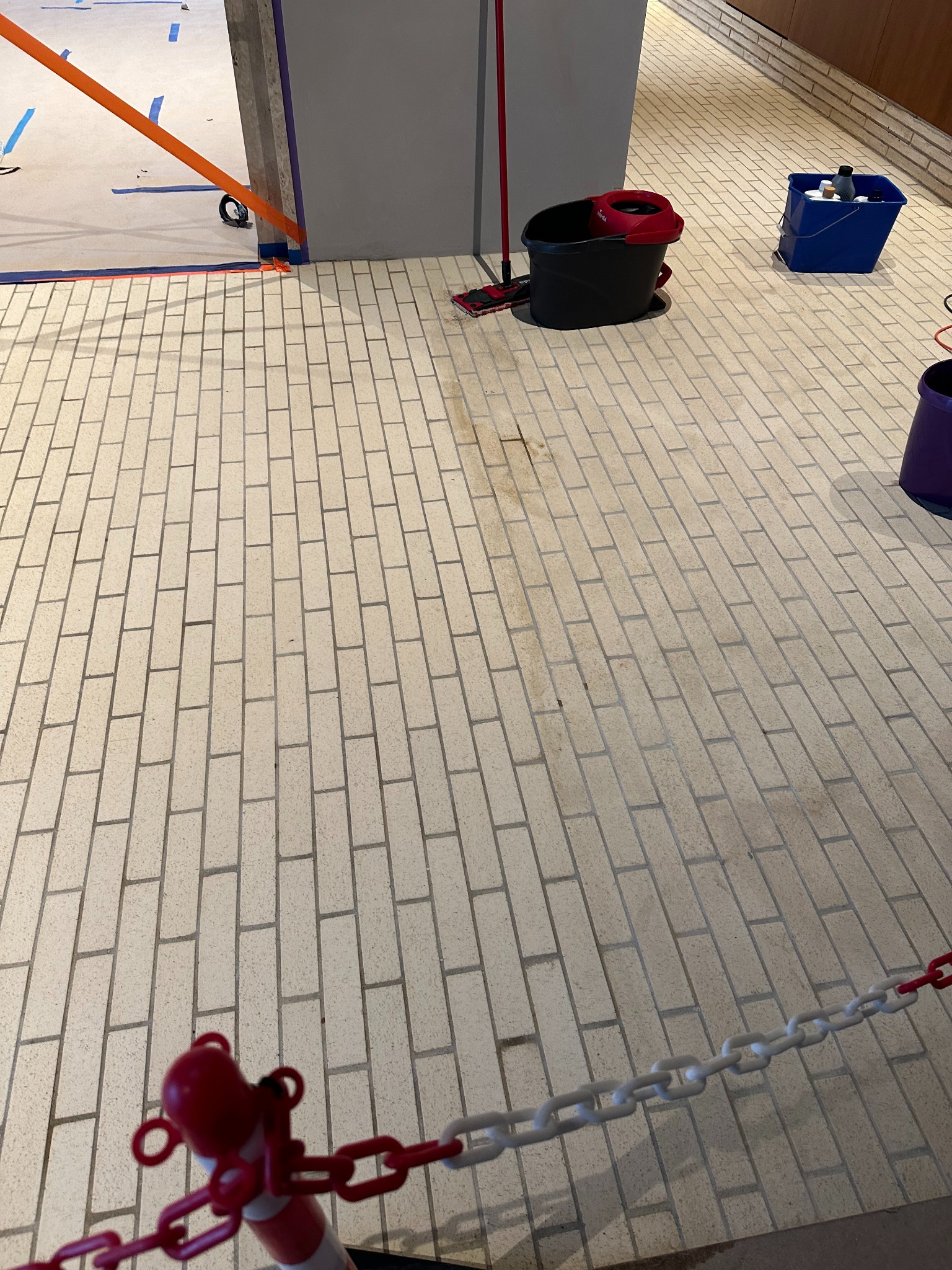 Podłoga z cegieł po remoncie, widoczne narzędzia i środki czystości, taśmy zabezpieczające. Fragment łańcucha oddzielającego strefę. Ślady po czyszczeniu.