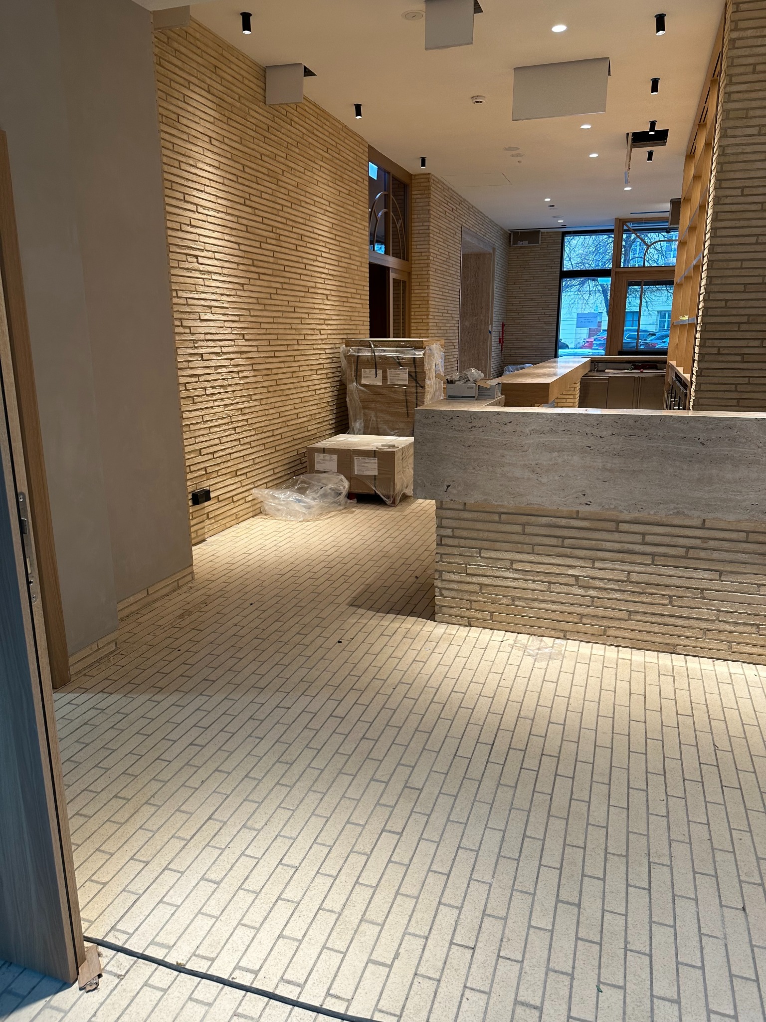 Wnętrze w trakcie wykańczania: ceglana ściana, podłoga z cegieł, widoczne kartony i elementy wyposażenia. Jasne oświetlenie, surowy, minimalistyczny styl.