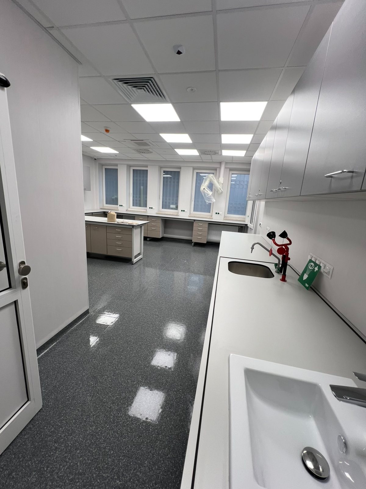 Wnętrze czystego, jasnego laboratorium z białymi szafkami, umywalkami i szarymi podłogami. Widoczne okna i sprzęt laboratoryjny. Minimalistyczny design.