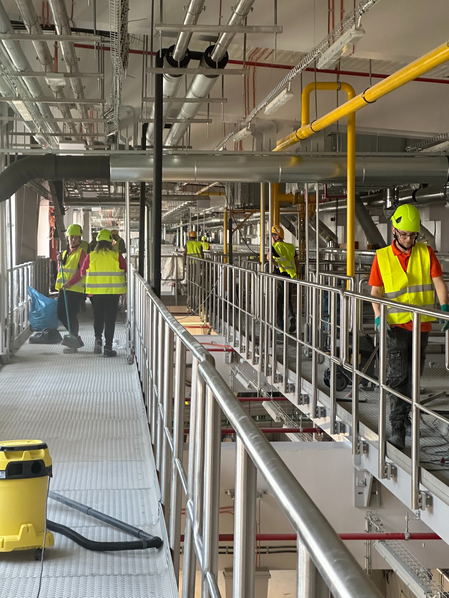 Ekipa sprzątająca w jaskrawożółtych kamizelkach i kaskach, z odkurzaczem i workiem na śmieci, na tle przemysłowej instalacji z rurami i kablami.