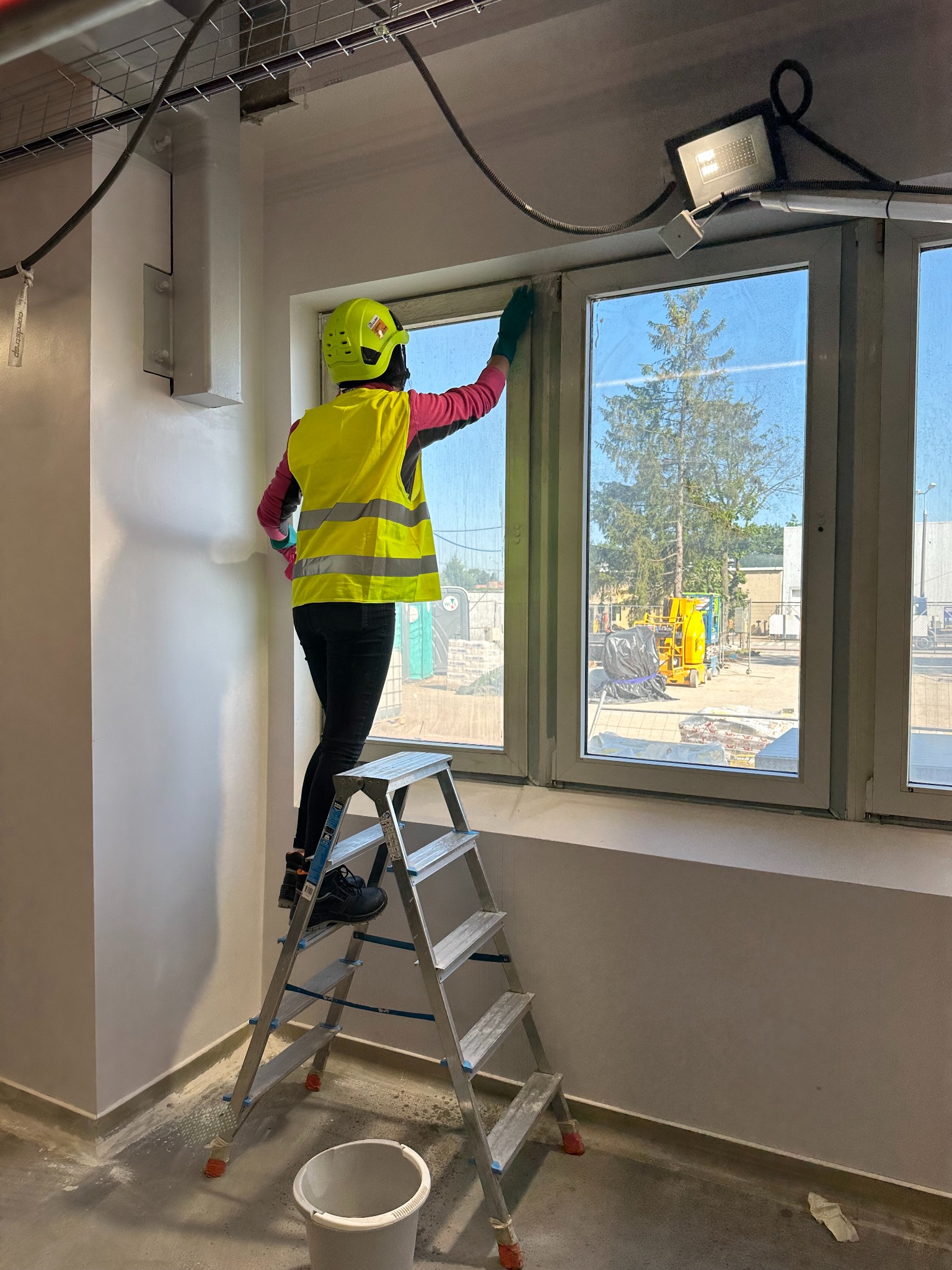 Pracownik w żółtej kamizelce i kasku myje okna na drabinie w nowym budynku. Widoczne przewody pod sufitem i sprzęt budowlany za oknem.