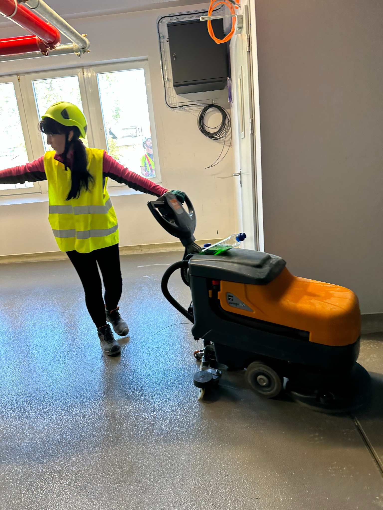 Pracownica w żółtej kamizelce i kasku obsługuje pomarańczową maszynę do czyszczenia podłóg w jasnym pomieszczeniu z oknem i instalacjami pod sufitem.