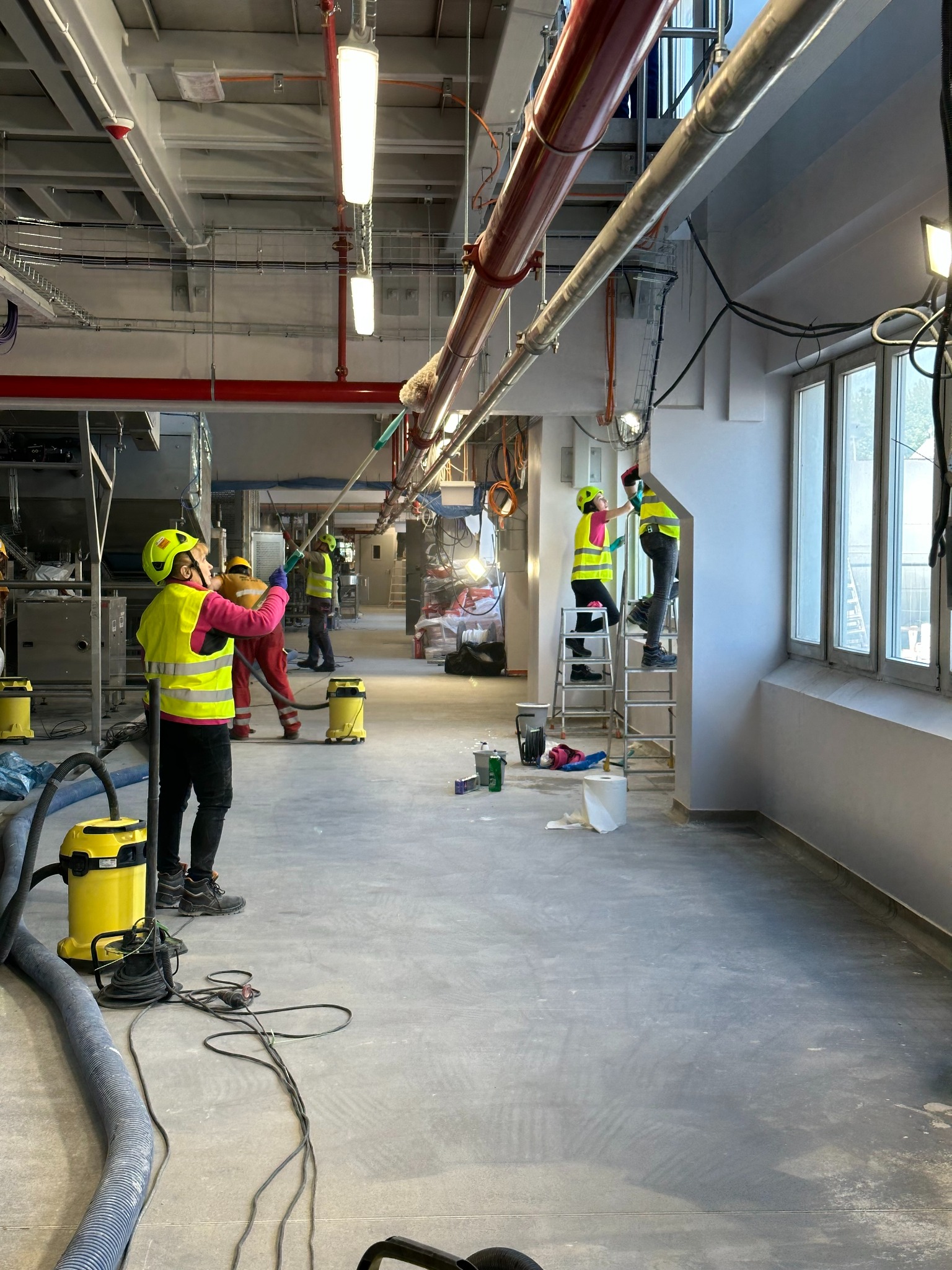 Ekipa w żółtych kamizelkach sprząta duże, przemysłowe pomieszczenie z oknami. Widoczne odkurzacze przemysłowe, drabina i inne narzędzia. Prace porządkowe w toku.