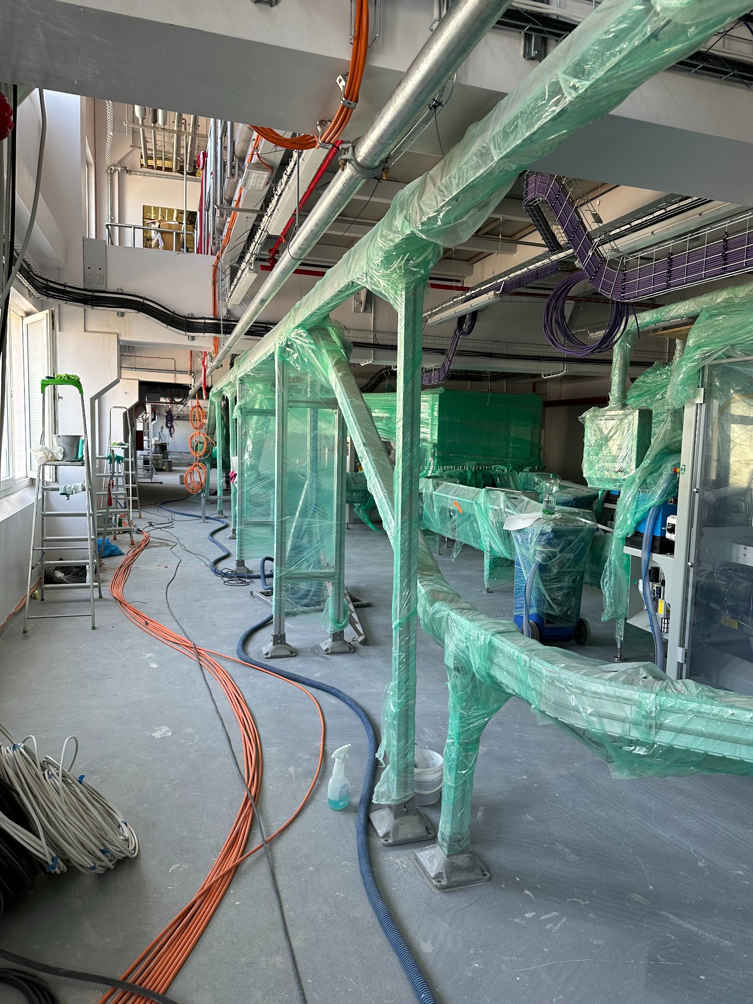 Wnętrze hali produkcyjnej w trakcie instalacji linii technologicznej. Elementy maszyn zabezpieczone folią, widoczne kable i rury. Etap montażu.