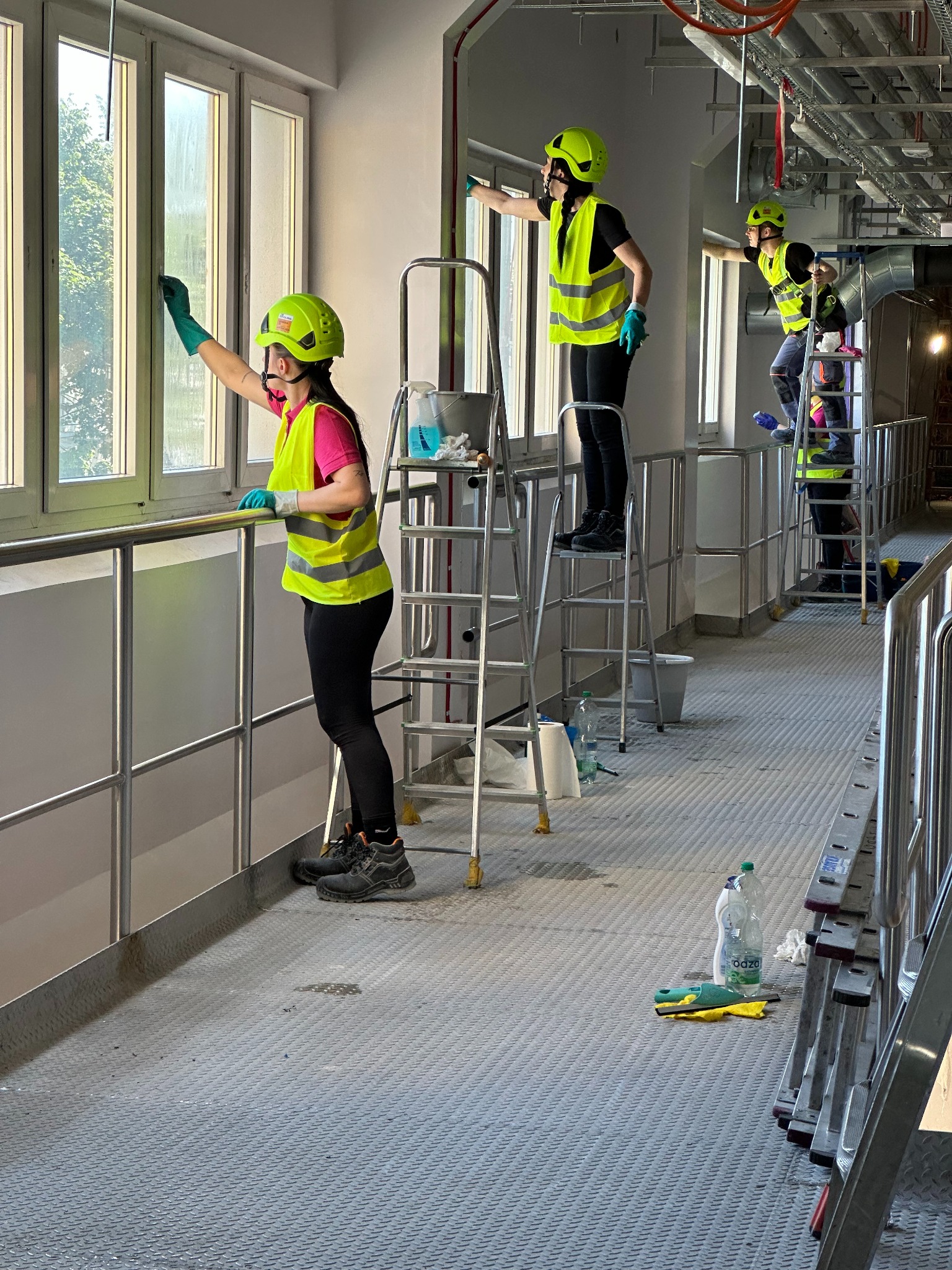 Ekipa w kaskach i kamizelkach na rusztowaniu myje okna w hali przemysłowej. Widoczne drabiny, środki czystości i narzędzia. Praca na wysokościach.
