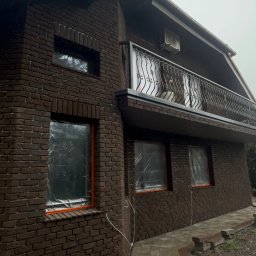 Pavlo Tsos - Dom z ciemnej cegły po renowacji elewacji, okna zabezpieczone folią, balkon z ozdobną balustradą. Widoczny fragment chodnika z kostki brukowej.