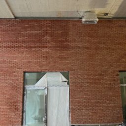 Pavlo Tsos - Ściana z cegły klinkierowej wewnątrz budynku, z oknami częściowo zakrytymi folią ochronną. Widoczne instalacje pod sufitem i fragment konstrukcji budynku.