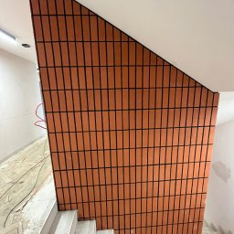 Pavlo Tsos - Ściana schodów wykończona ceglastymi płytkami z ciemną fugą, widok z dołu. Schody w trakcie prac wykończeniowych, widoczne kable i zabrudzenia.