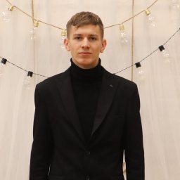 Pavlo Tsos - Młody mężczyzna w czarnym golfie i marynarce, na tle białej zasłony i girlandy z żarówek. Portret w neutralnym oświetleniu, z delikatnym uśmiechem.