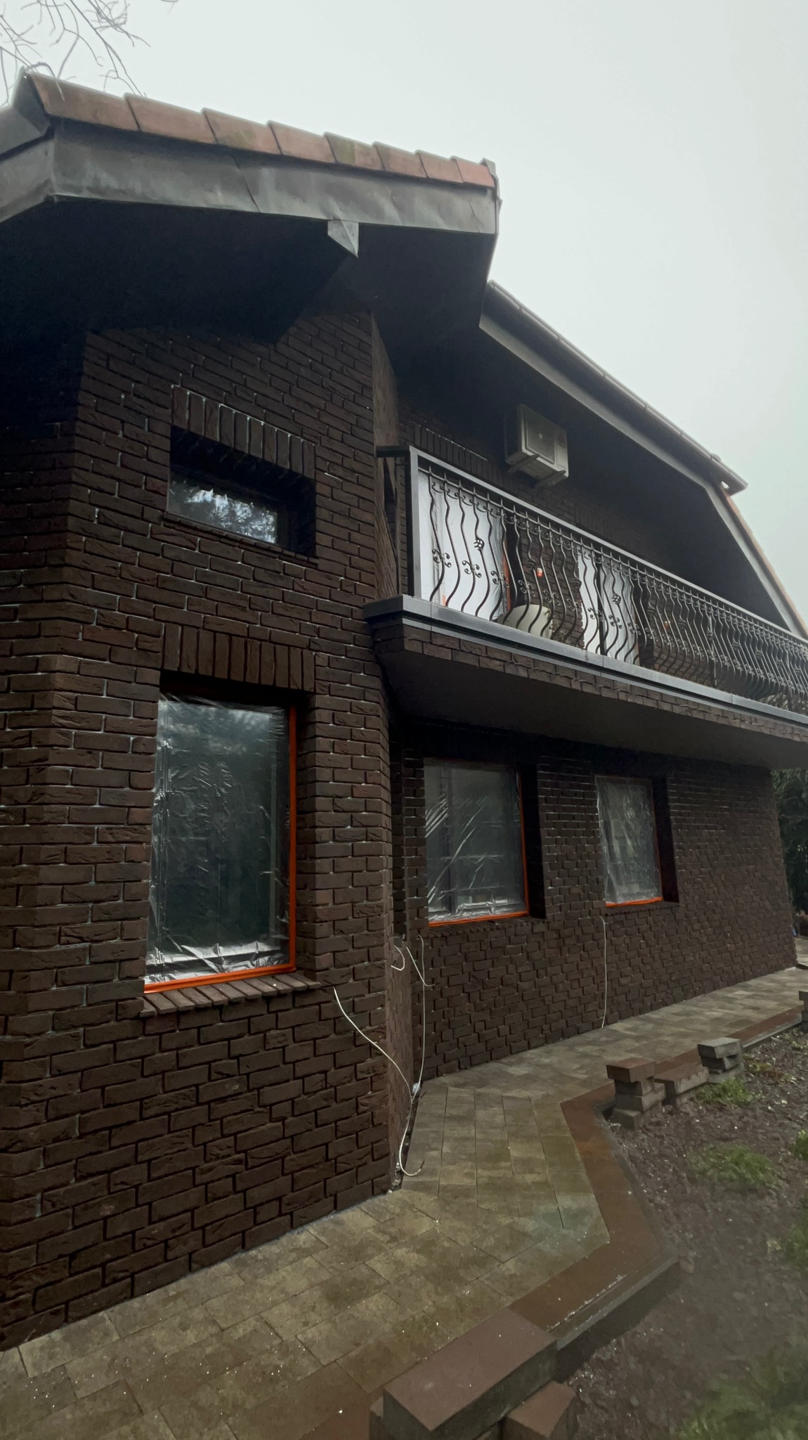 Dom z ciemnej cegły po renowacji elewacji, okna zabezpieczone folią, balkon z ozdobną balustradą. Widoczny fragment chodnika z kostki brukowej.