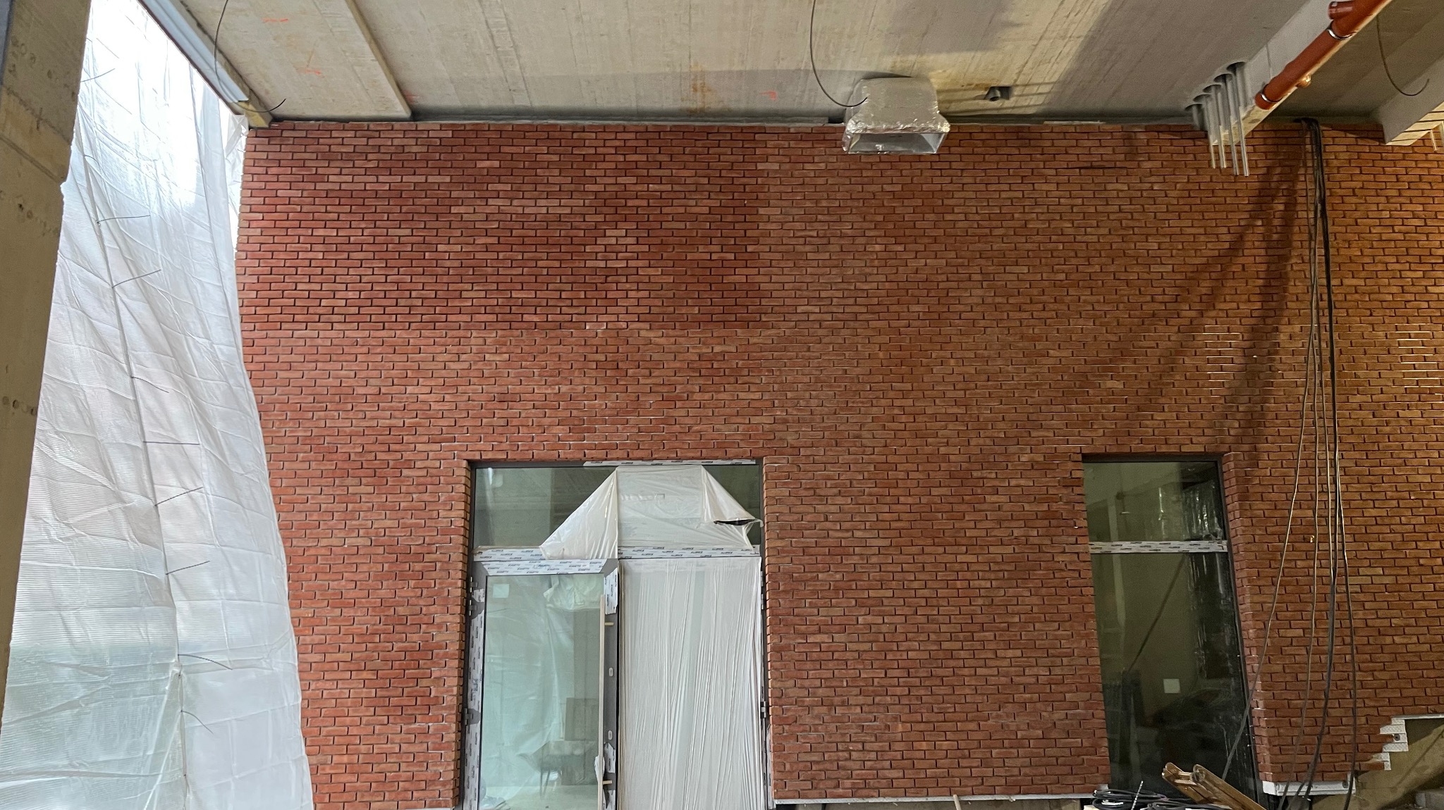 Ściana z cegły klinkierowej wewnątrz budynku, z oknami częściowo zakrytymi folią ochronną. Widoczne instalacje pod sufitem i fragment konstrukcji budynku.