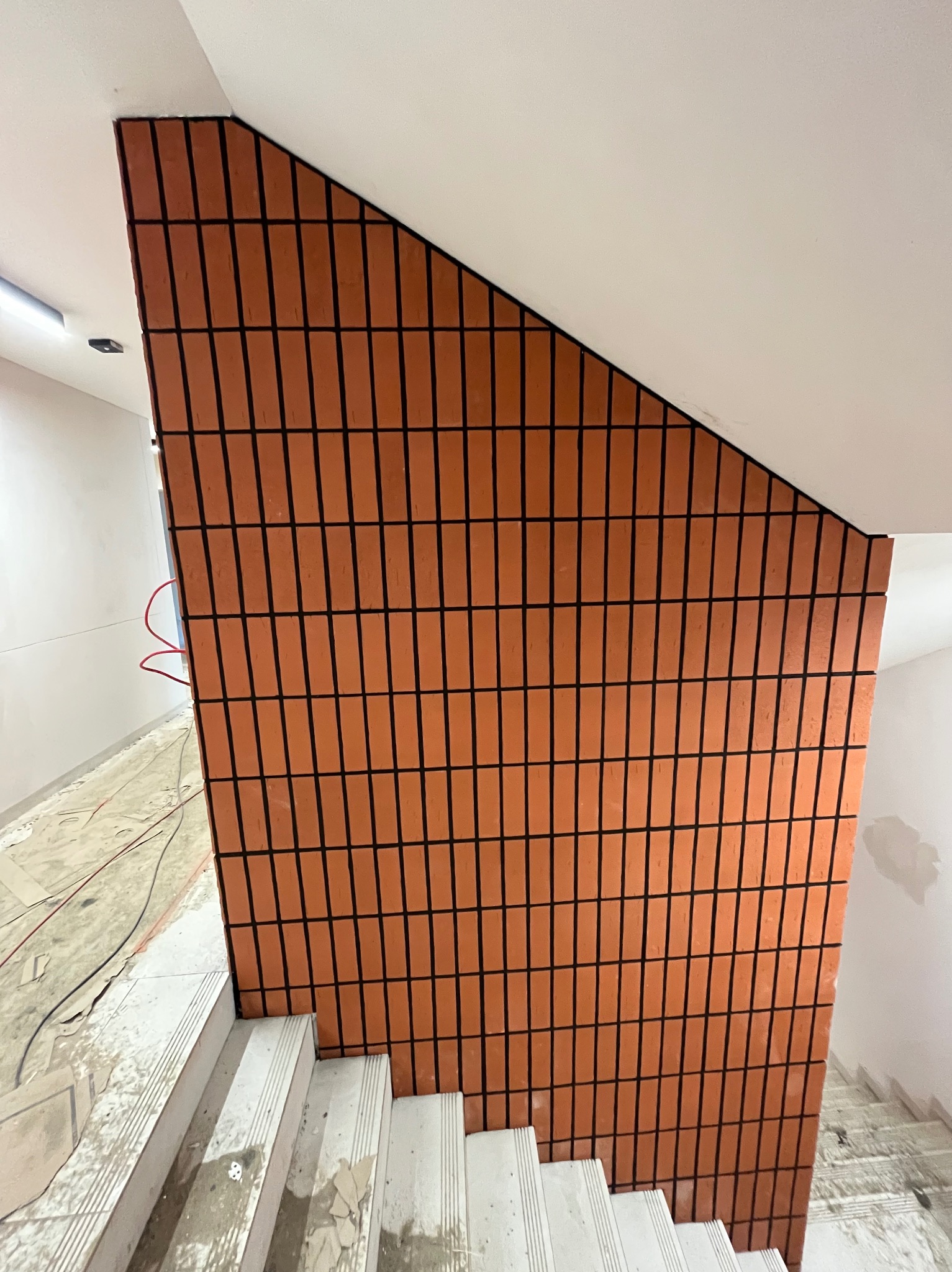 Ściana schodów wykończona ceglastymi płytkami z ciemną fugą, widok z dołu. Schody w trakcie prac wykończeniowych, widoczne kable i zabrudzenia.