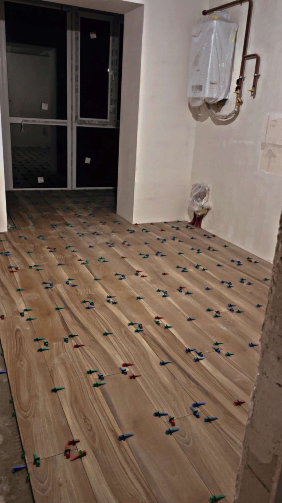 Układanie drewnopodobnych płytek podłogowych z użyciem klipsów poziomujących w jasnym wnętrzu. Widoczny fragment ściany z piecem gazowym w folii.