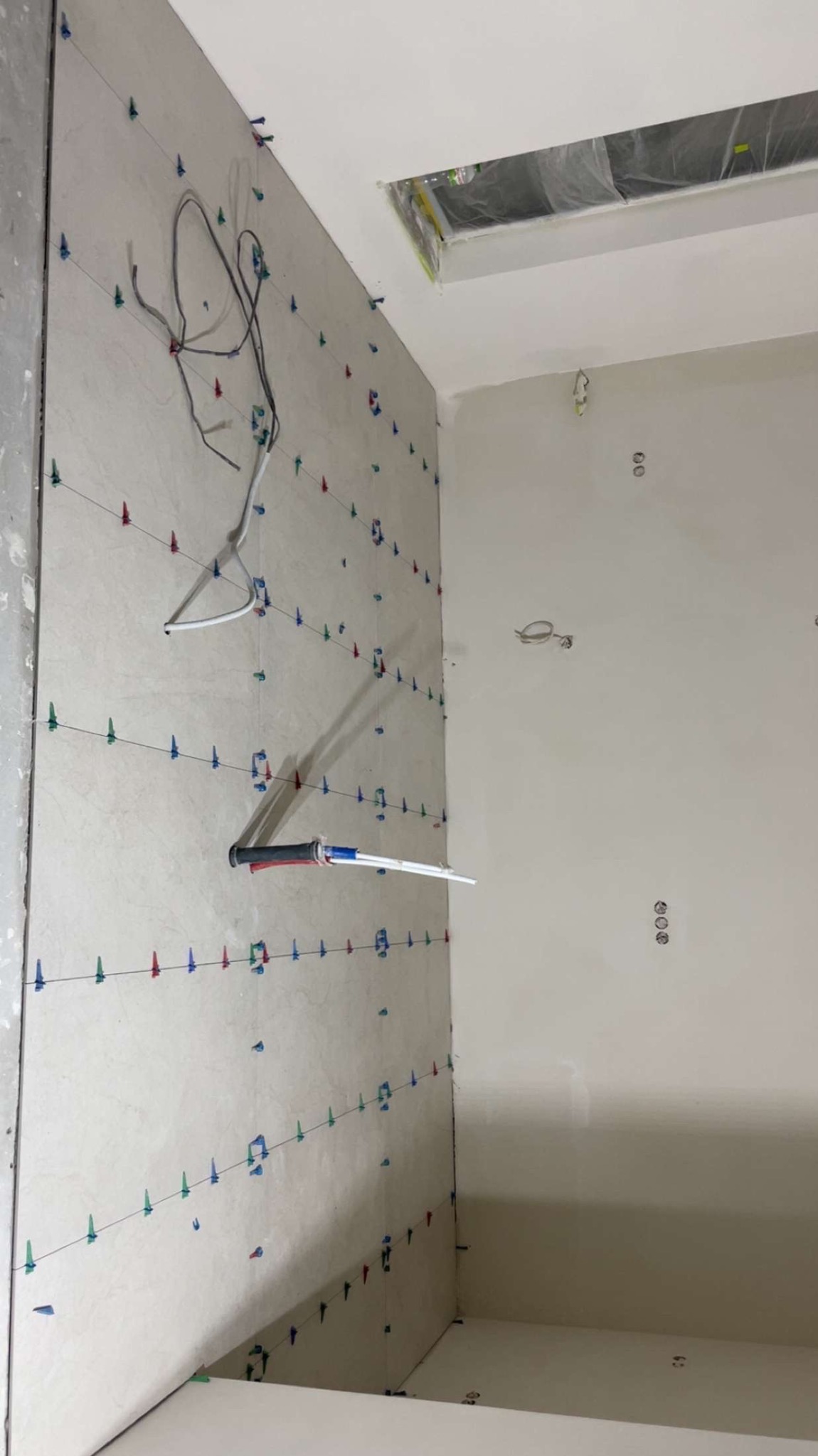 Ściana w trakcie układania płytek, widoczne kolorowe kliny poziomujące i narzędzie do aplikacji. Częściowo wykończona powierzchnia, surowy stan.