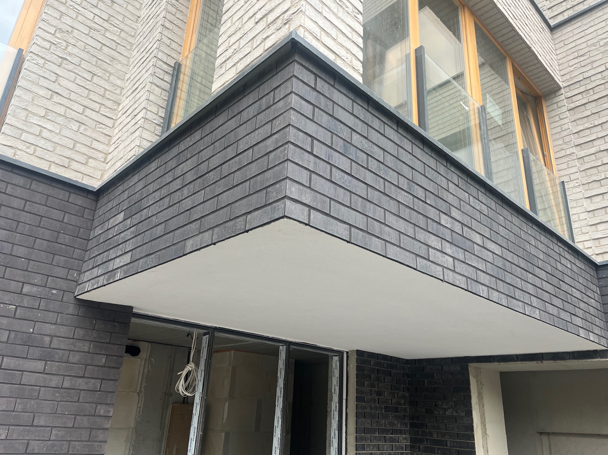 Wykończona elewacja budynku z szarej cegły klinkierowej, z widocznym fragmentem balkonu z szklaną balustradą i oknami. Detal architektoniczny.