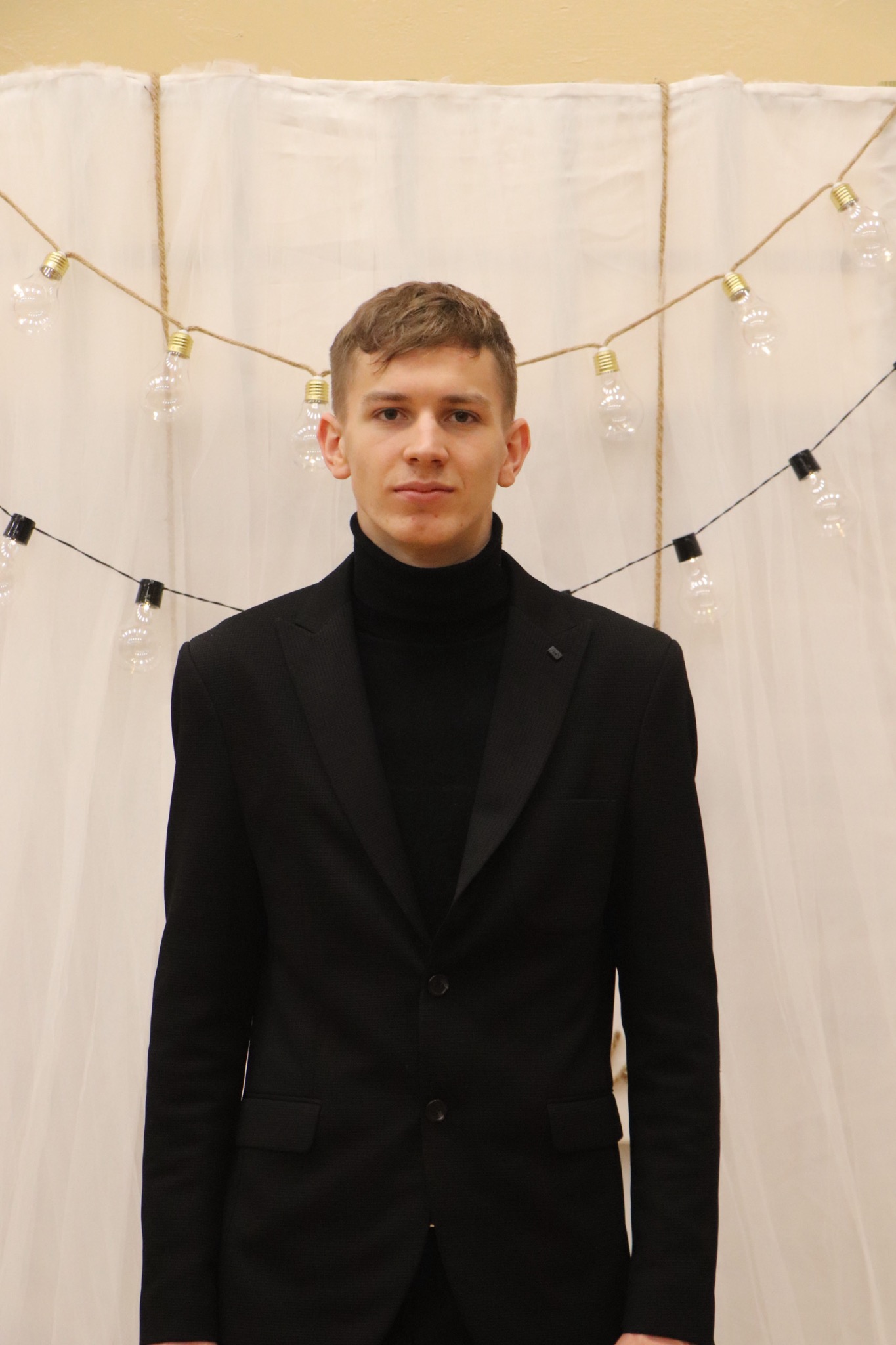 Młody mężczyzna w czarnym golfie i marynarce, na tle białej zasłony i girlandy z żarówek. Portret w neutralnym oświetleniu, z delikatnym uśmiechem.
