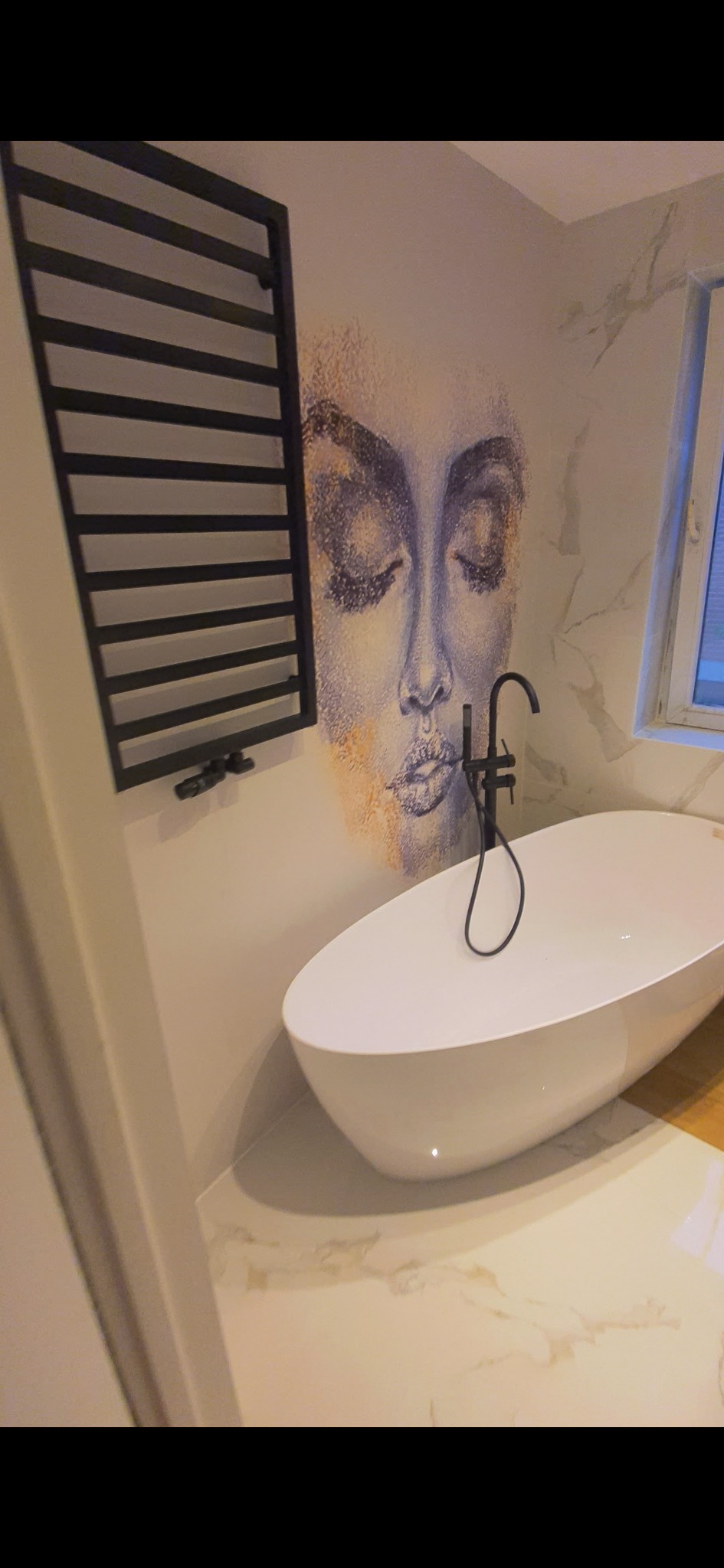 Nowoczesna łazienka z wolnostojącą wanną i czarną armaturą. Na ścianie dekoracyjny rysunek twarzy i grzejnik drabinkowy w czarnym kolorze.