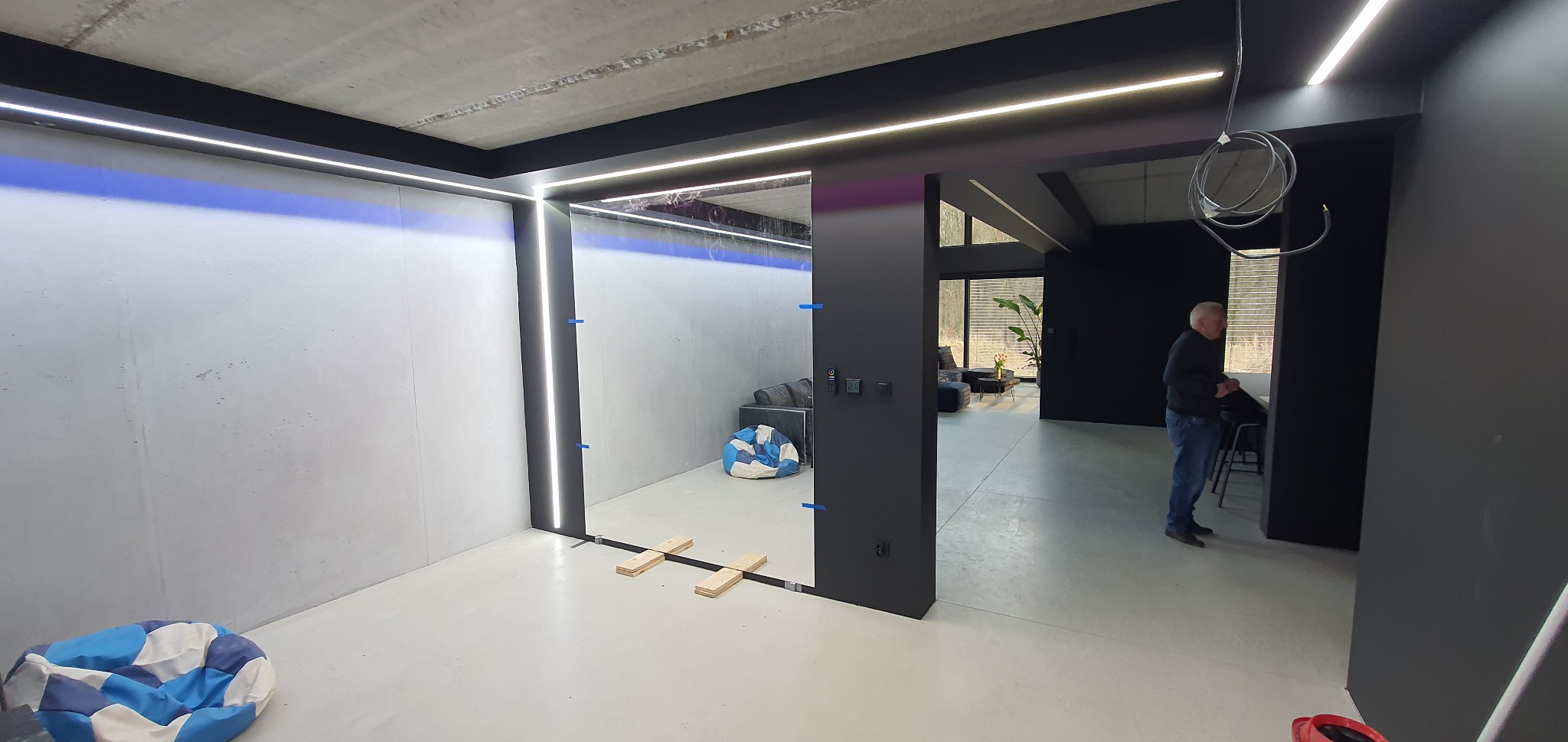 Nowoczesne wnętrze z przesuwnymi drzwiami i oświetleniem LED. Widoczny fragment salonu i osoba w tle. Minimalistyczny design z betonowymi ścianami i czarnymi elementami.