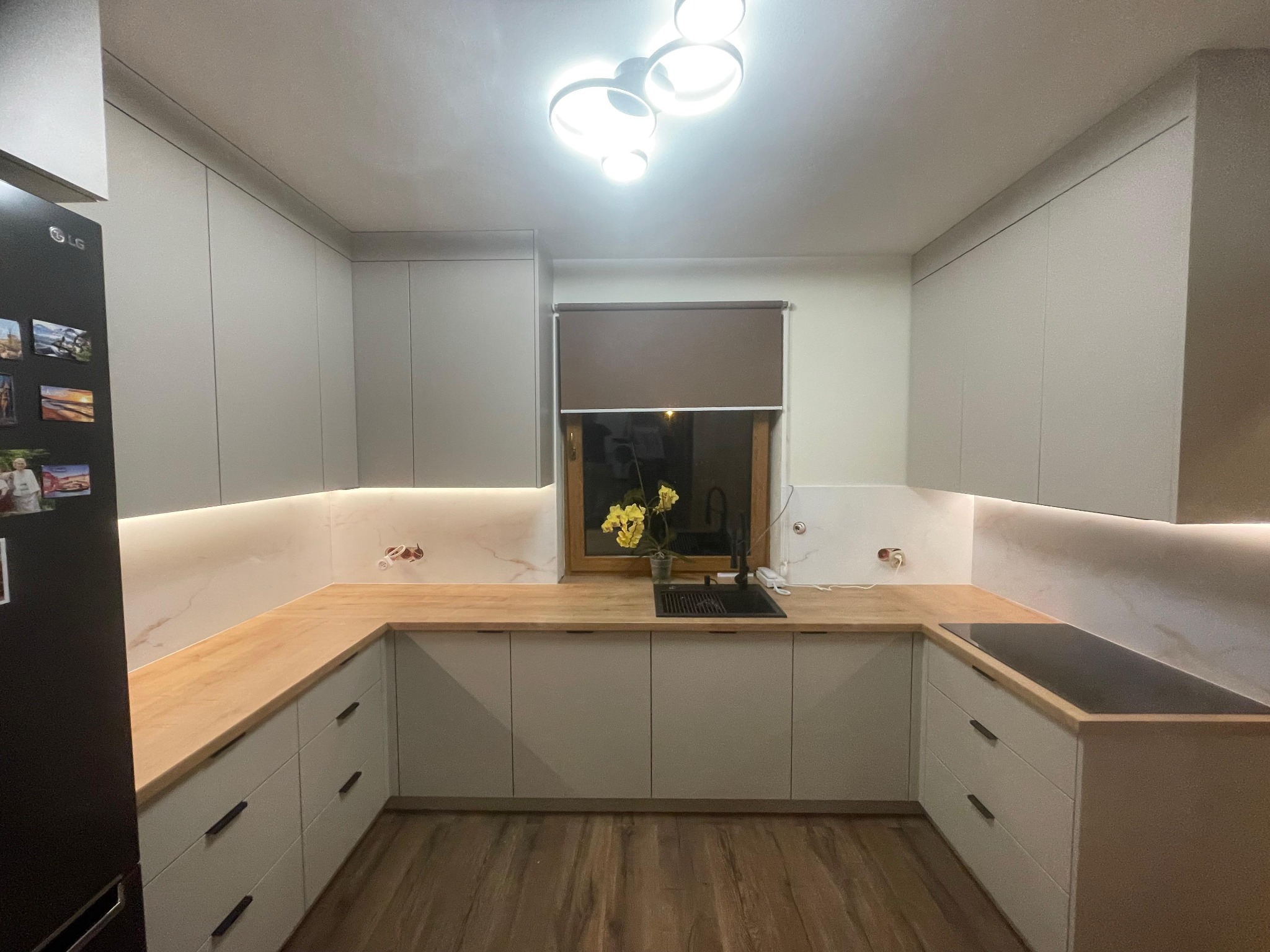 Nowoczesna kuchnia w odcieniach szarości z drewnianym blatem, oświetleniem LED pod szafkami i kwiatami w oknie. Minimalistyczny design z czarną armaturą i płytą indukcyjną.