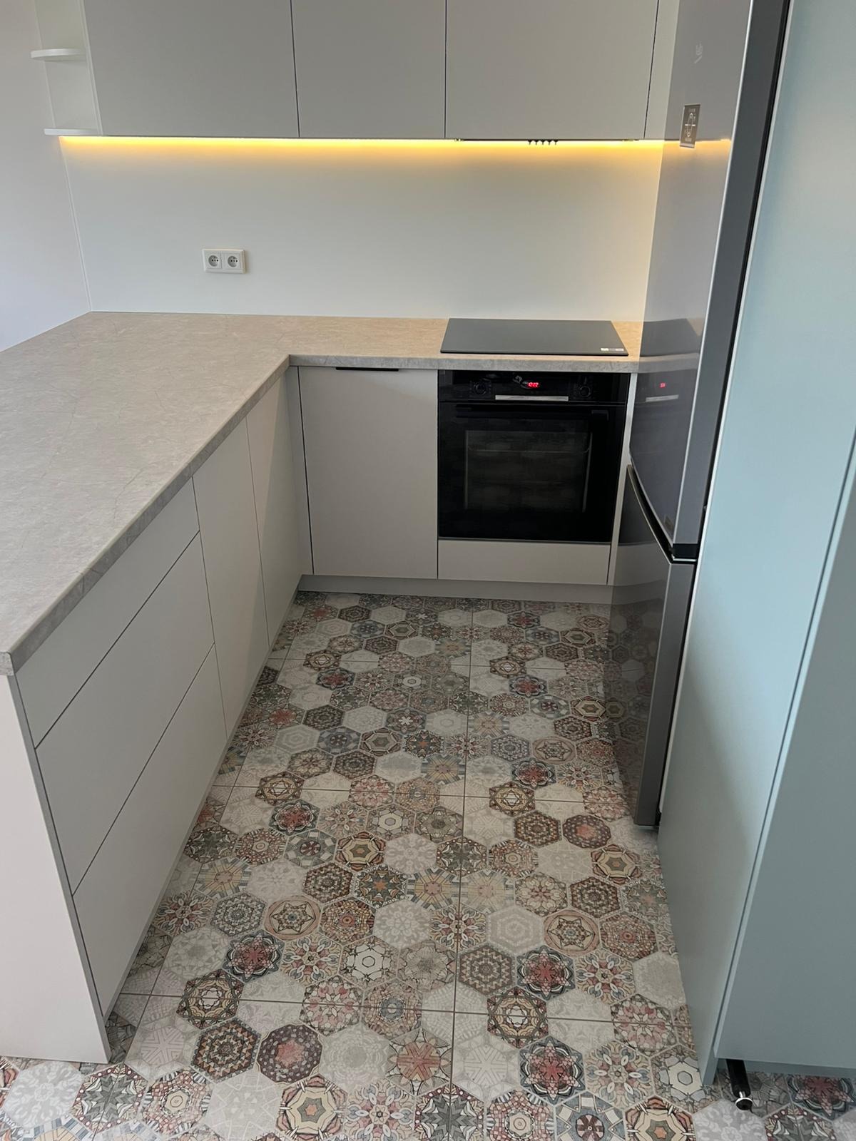 Jasna kuchnia z heksagonalną mozaiką podłogową. Minimalistyczne szafki, blat z subtelnym wzorem i oświetlenie LED pod szafkami górnymi. Nowoczesny piekarnik i lodówka.