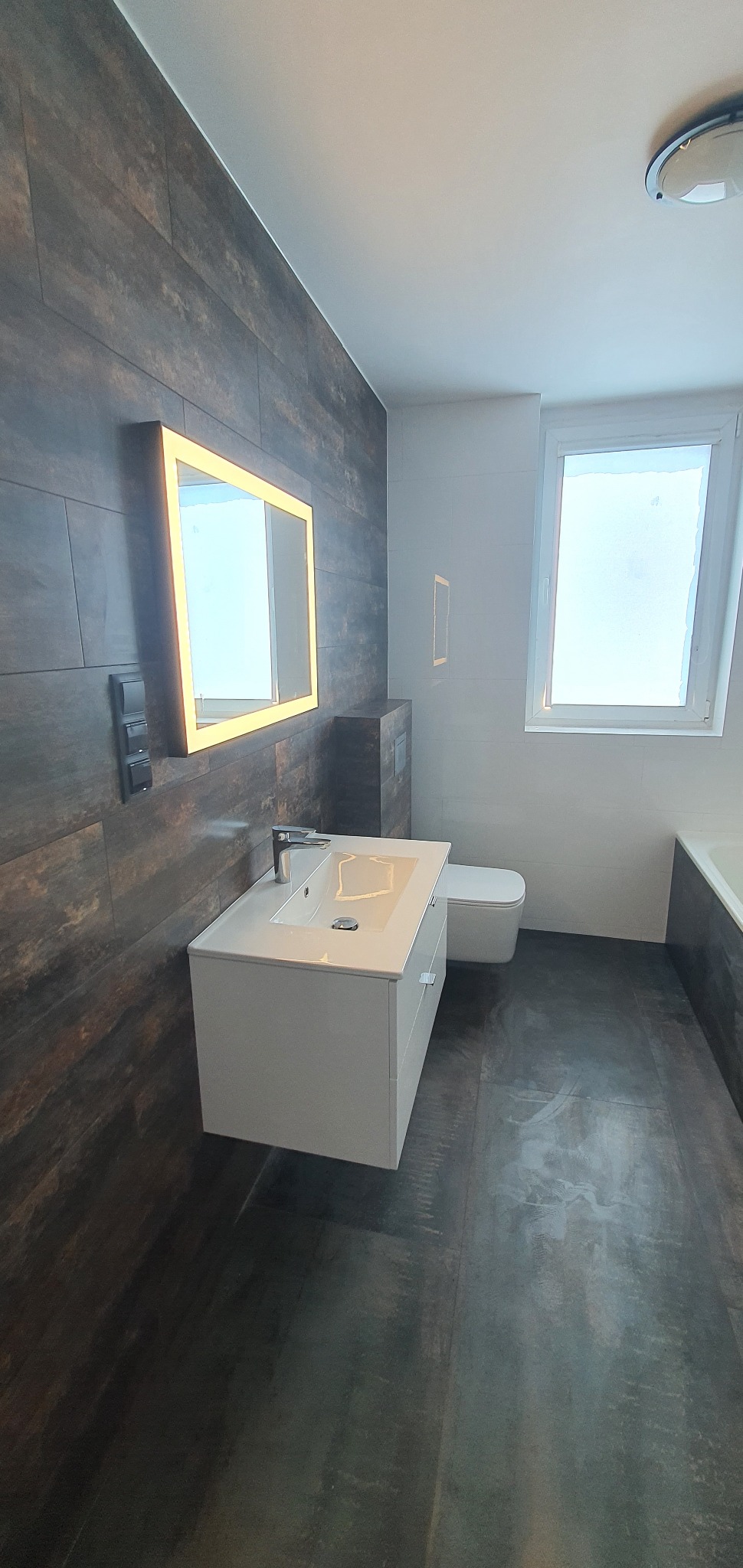 Nowoczesna łazienka w Łodzi z podświetlanym lustrem, ciemnymi płytkami imitującymi drewno na ścianie i podłodze, białą umywalką z szafką i toaletą.