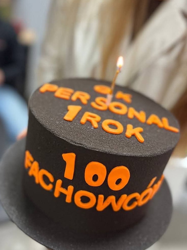 Czarny tort z pomarańczowymi napisami '1 ROK PERSONAL' i '100 FACHOWCÓW', zwieńczony płonącą świeczką, celebrujący rocznicę lub osiągnięcie firmy.
