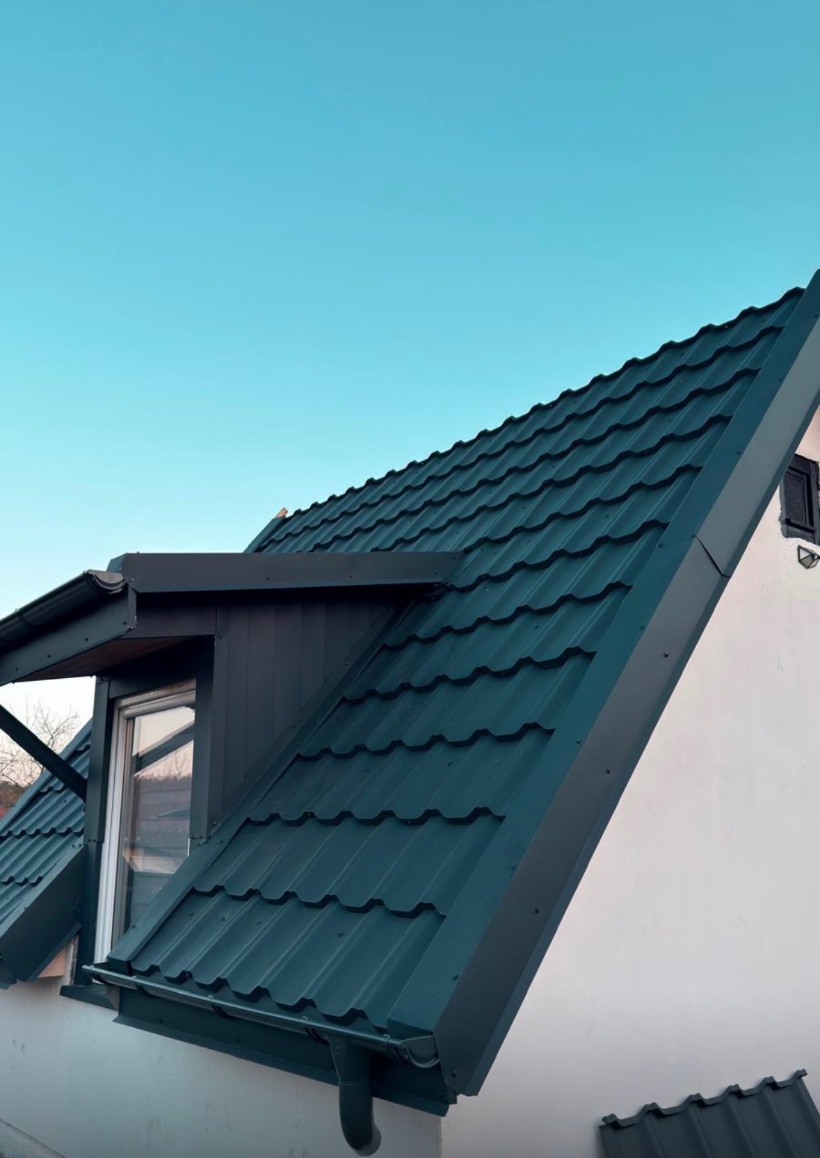 Nowy, ciemnozielony dach z blachy trapezowej na białym domu z oknem dachowym i rynną, widziany pod kątem. Czyste niebo w tle. Detal obróbki blacharskiej.