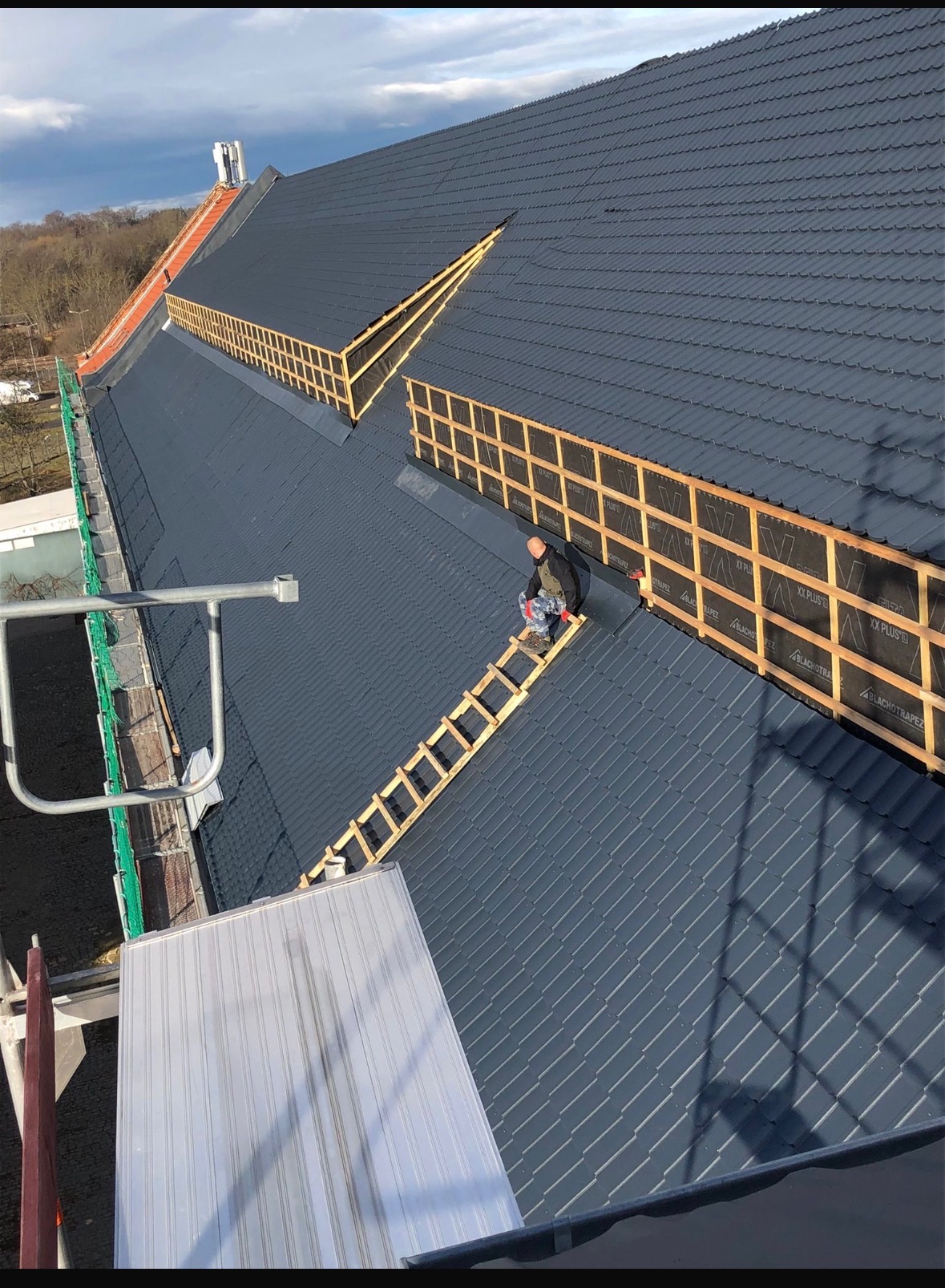 Dekarz na dachu z drabiny, montuje folię dachową. Widok z góry na szary dach z drewnianą konstrukcją i folią. W tle drzewa i niebo.